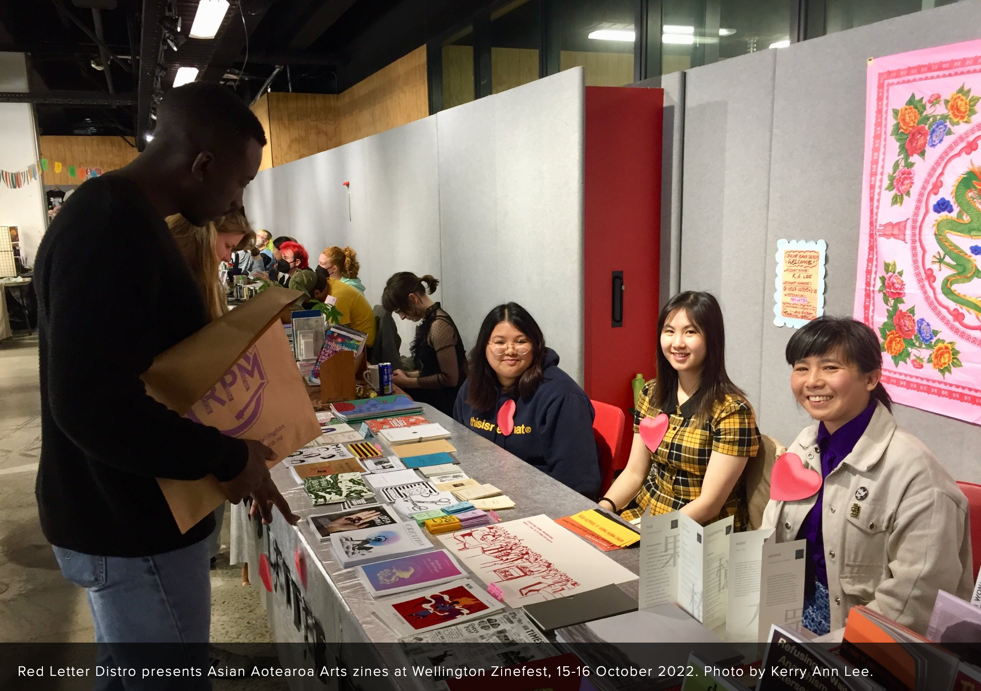 Three people smiling while manning a zine table and customer browsers the zines.