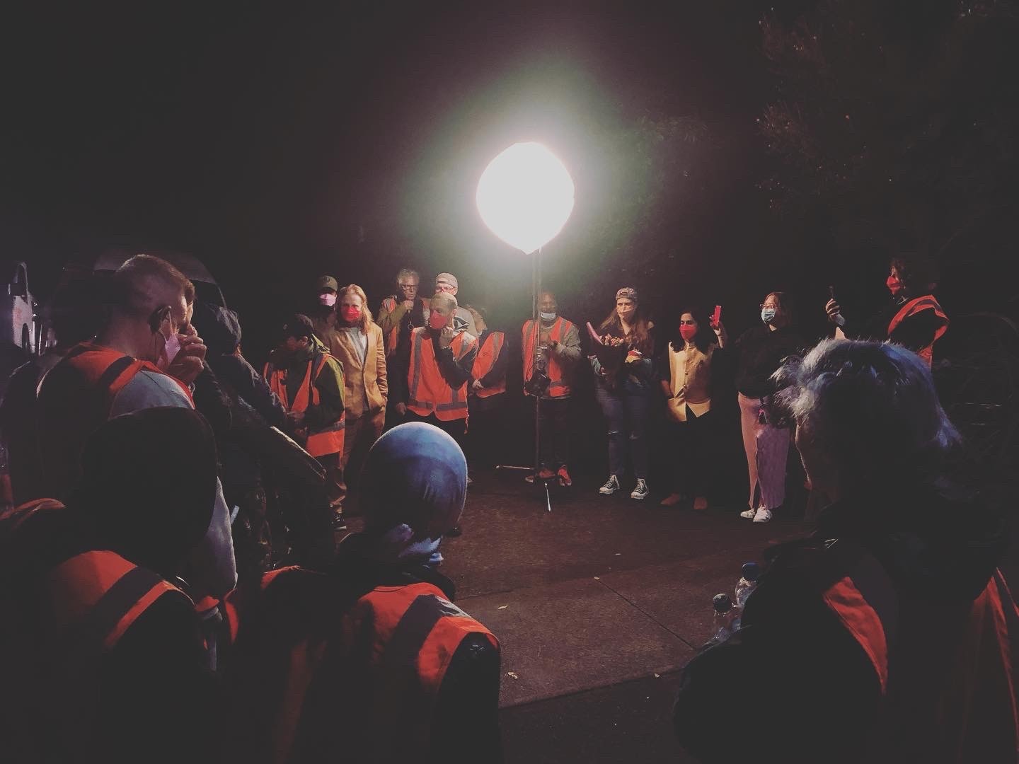 A film crew gathers in a circle at night under a bright light, many wearing orange safety vests and face masks.
