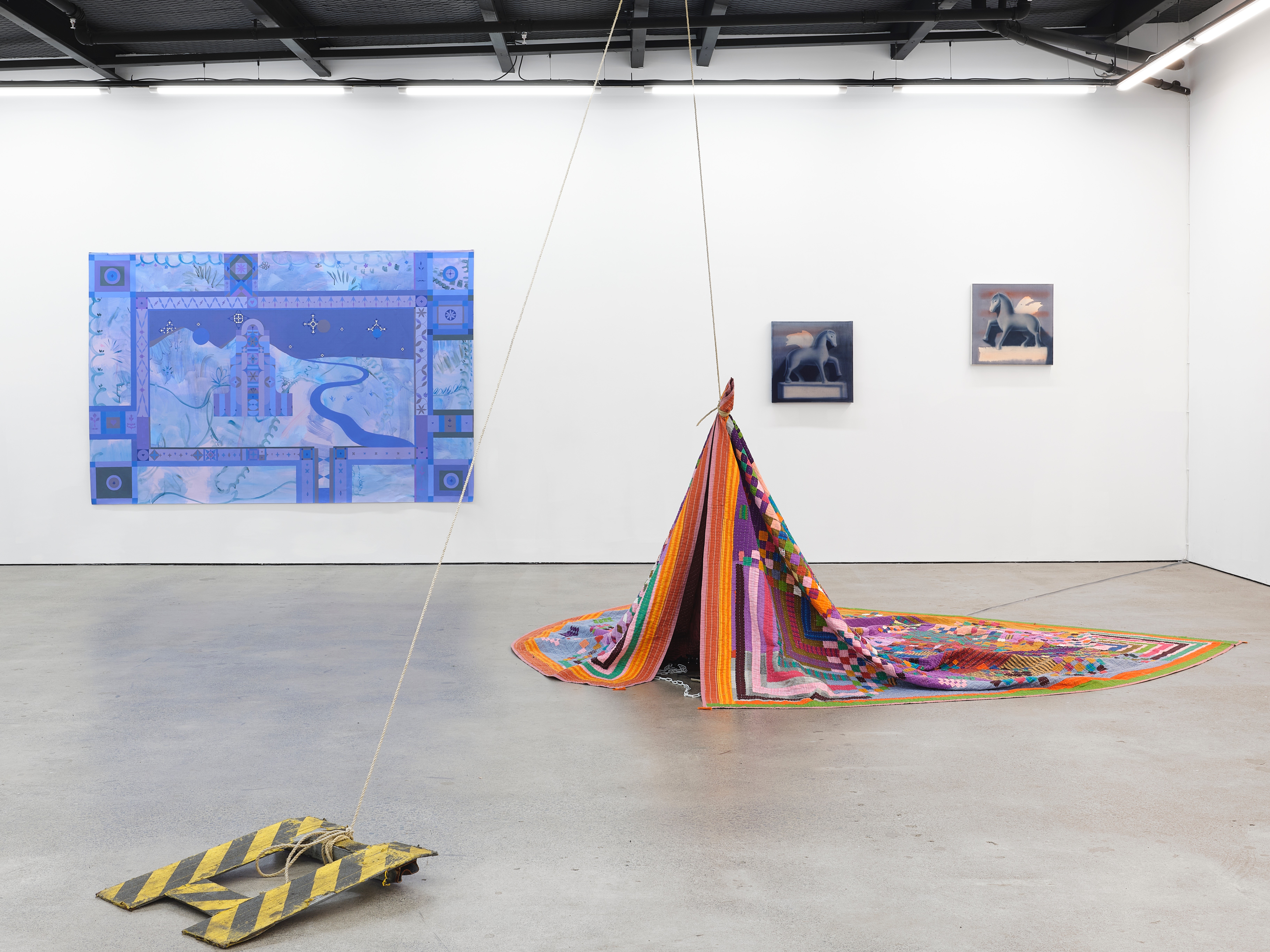 Photograph of an art exhibition. There are paintings on a white wall and on the grey floor is a installation of a quilt and small black and yellow industrial ramp.