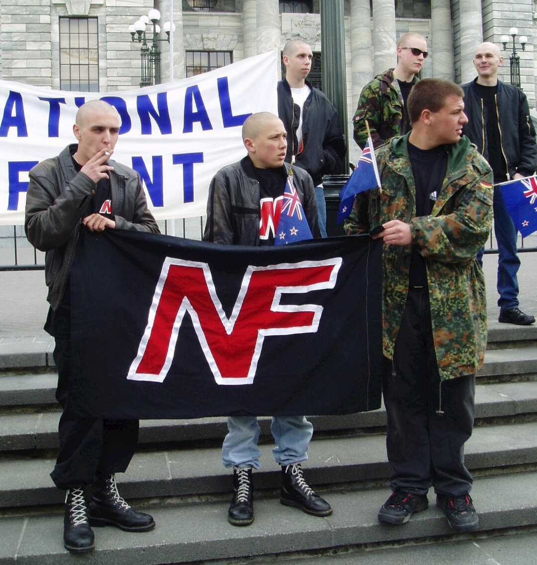 Skinheads with bald heads and combat boots hold a National Front flag in front of NZ Parliament.