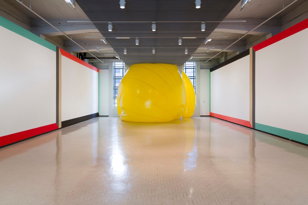 Large inflated yellow orb sandwiched between the floor and ceiling in a gallery space with minimal, frame-like wall paintings in red, green, gold and black on the walls either side. 