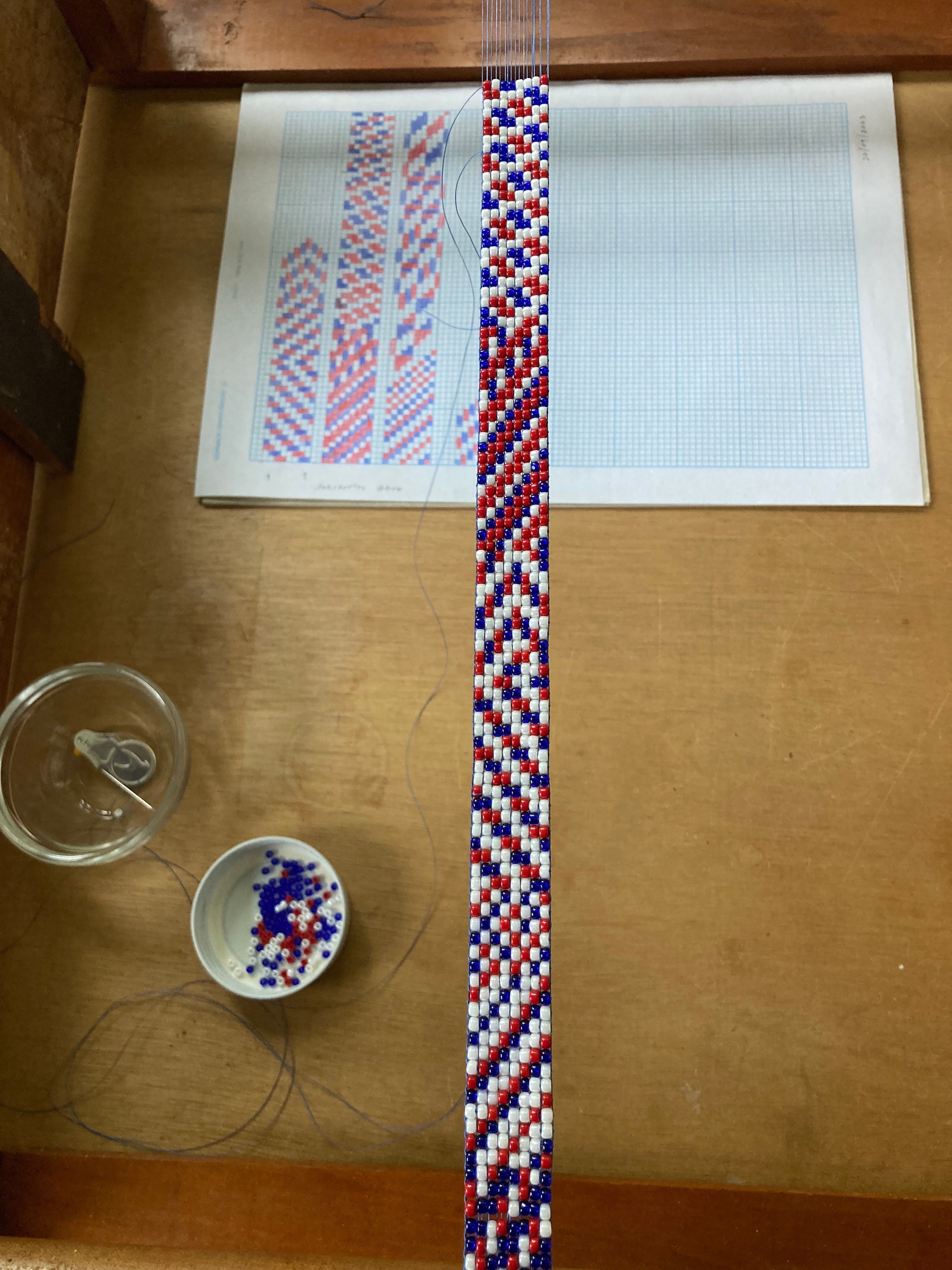 A red, white and blue beaded strap lies over a plan for the strap sketched on graph paper, next to two bowls of beads.