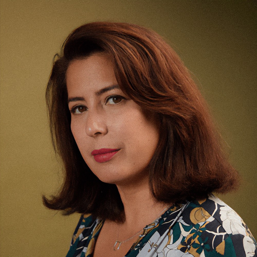 A woman with shoulder-length brown hair and red lipstick looks at the camera against an olive background.