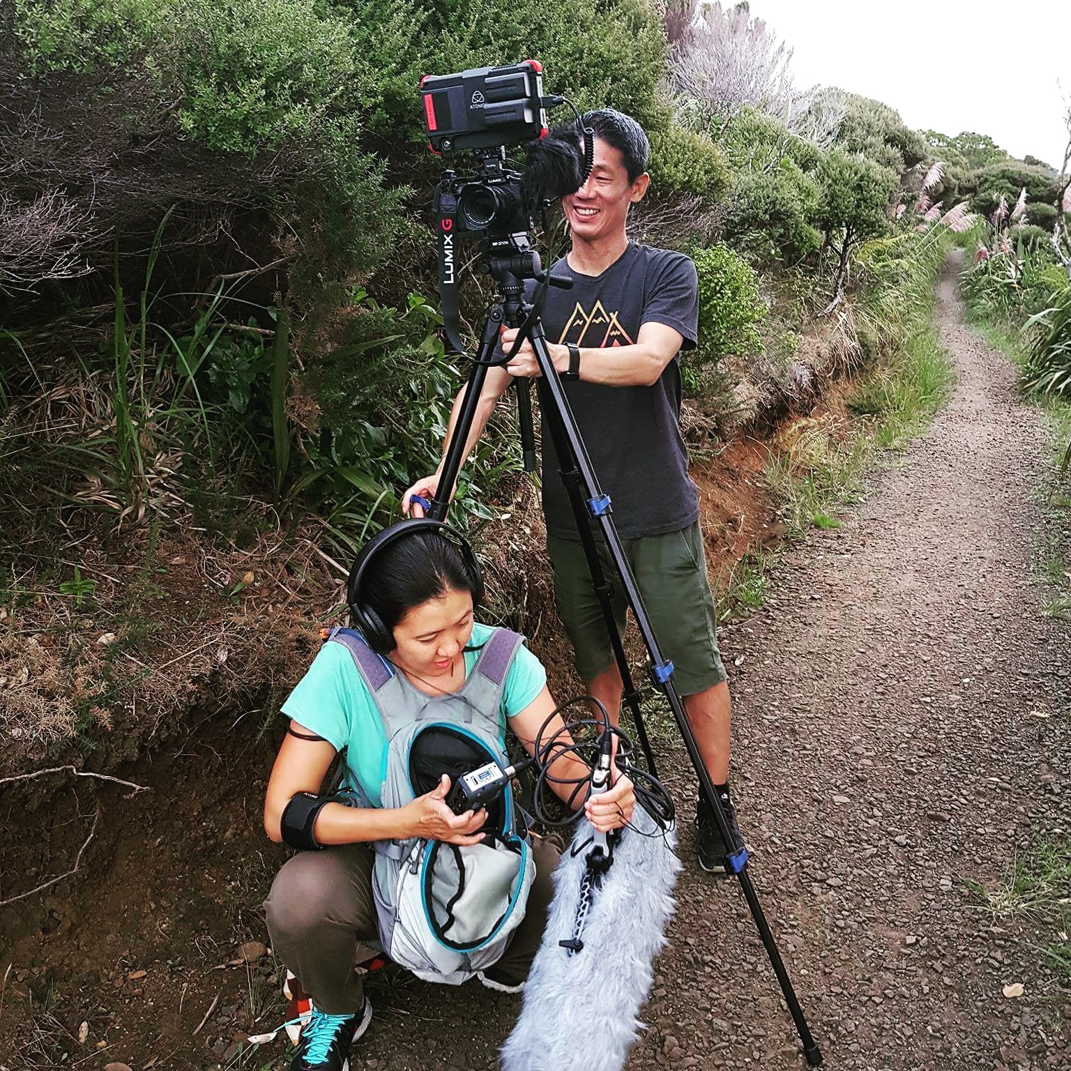 During a film shoot, a woman operating sound equipment is crouched in front of a video camera on a tripod, operating by a man standing behind it.