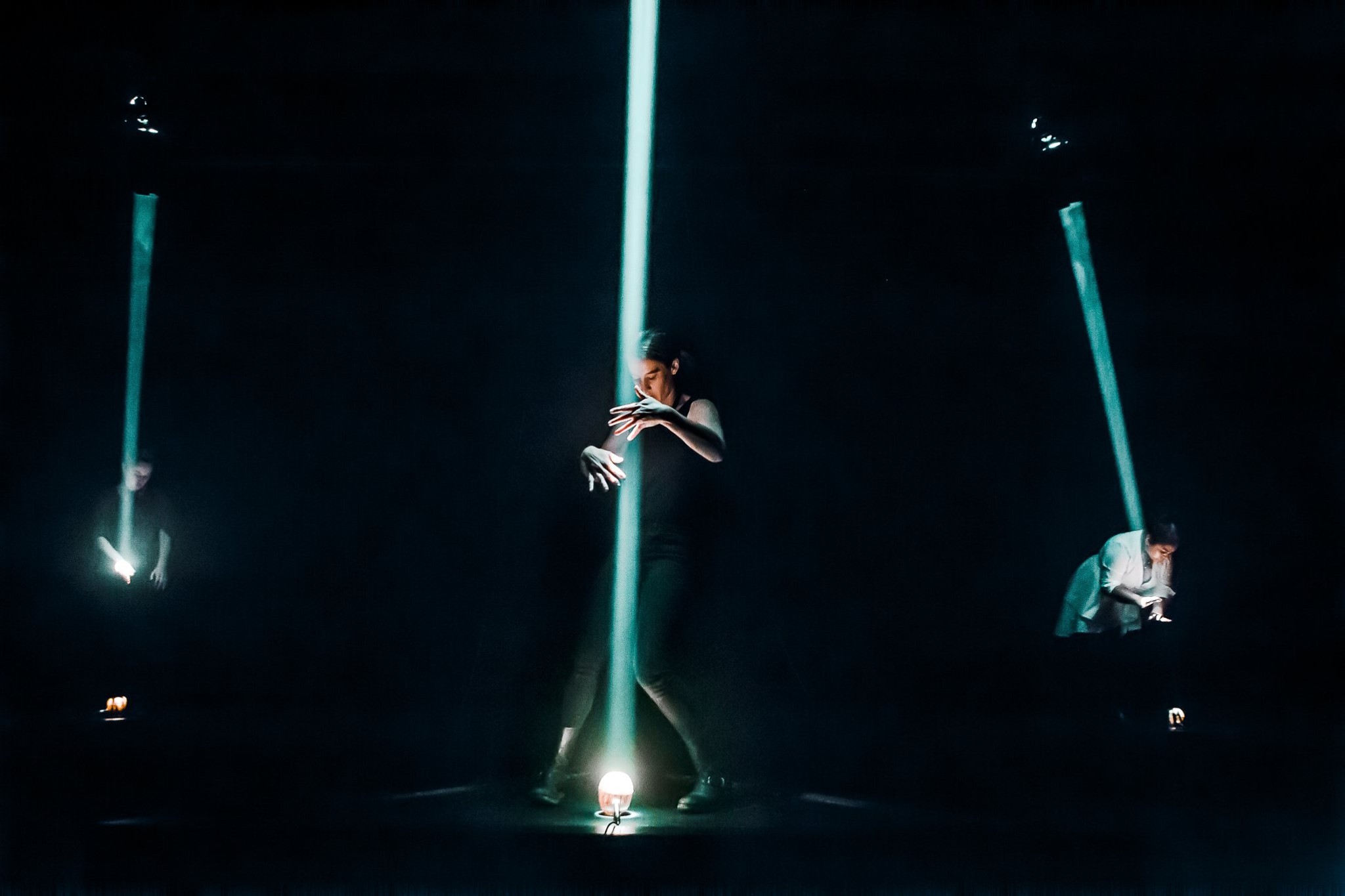 three women standing in a triangle, each lit by a thin, sharp green beam of light emanating from the ground