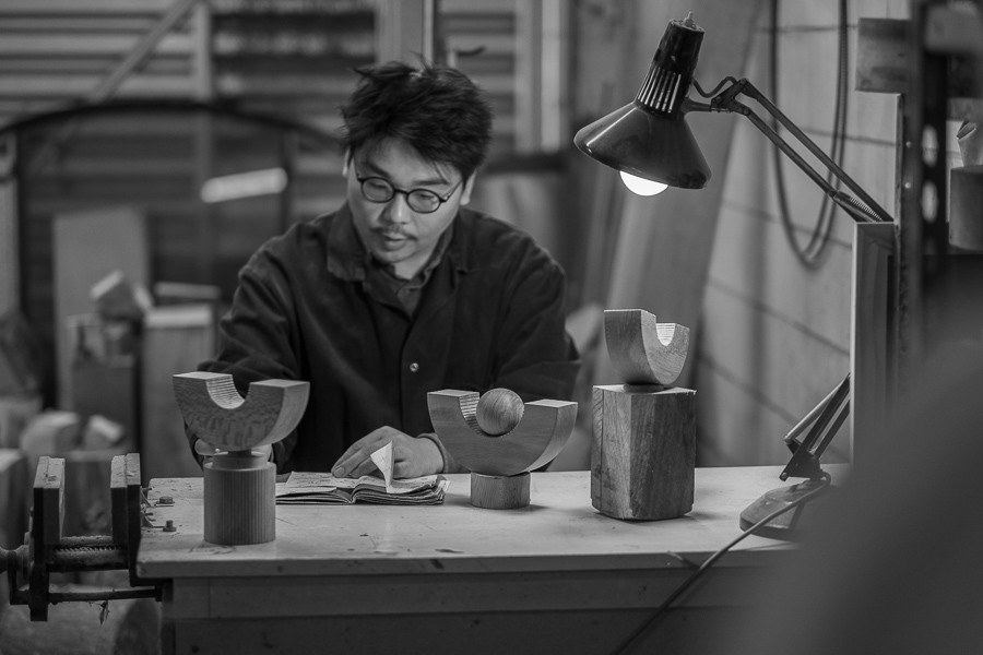 A black and white photo of a man with black hair and glasses working at a desk, flicking through his notebook while surrounded by wooden creations