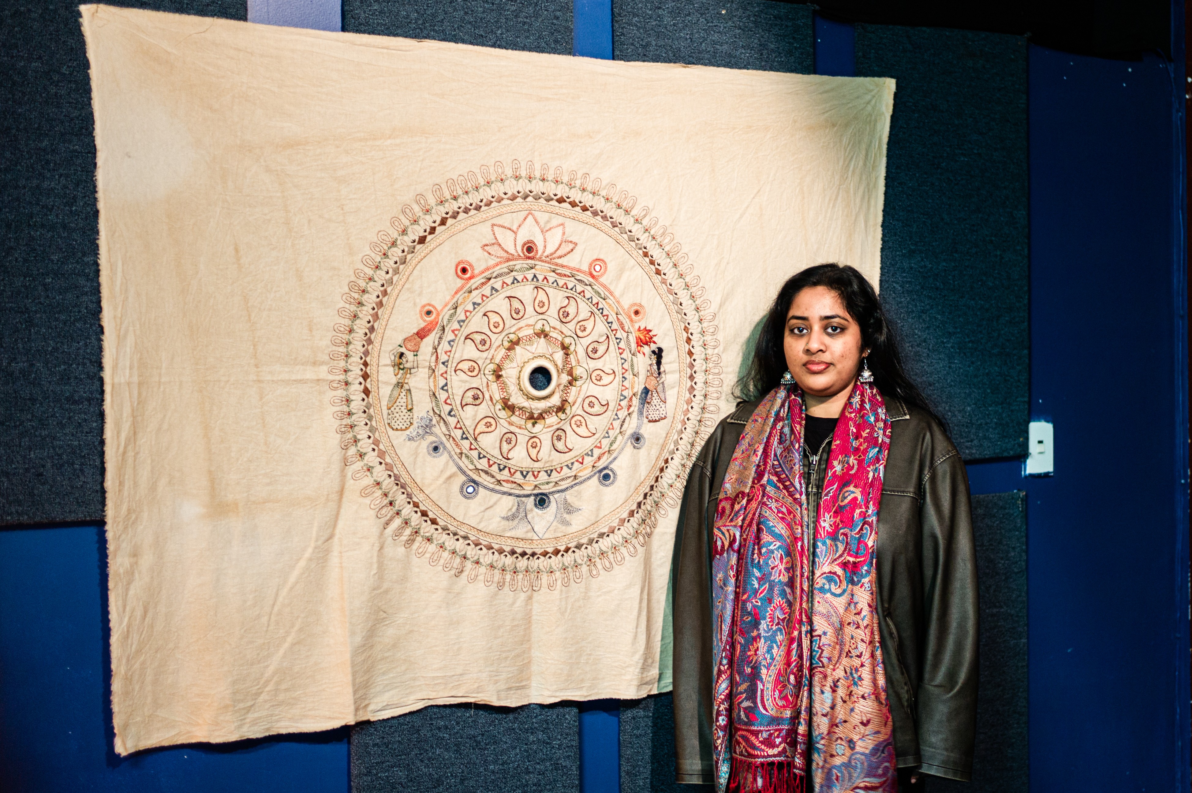 A woman with long dark hair standing beside a large piece of fabric with a circular embroidered pattern, hanging from the wall. 