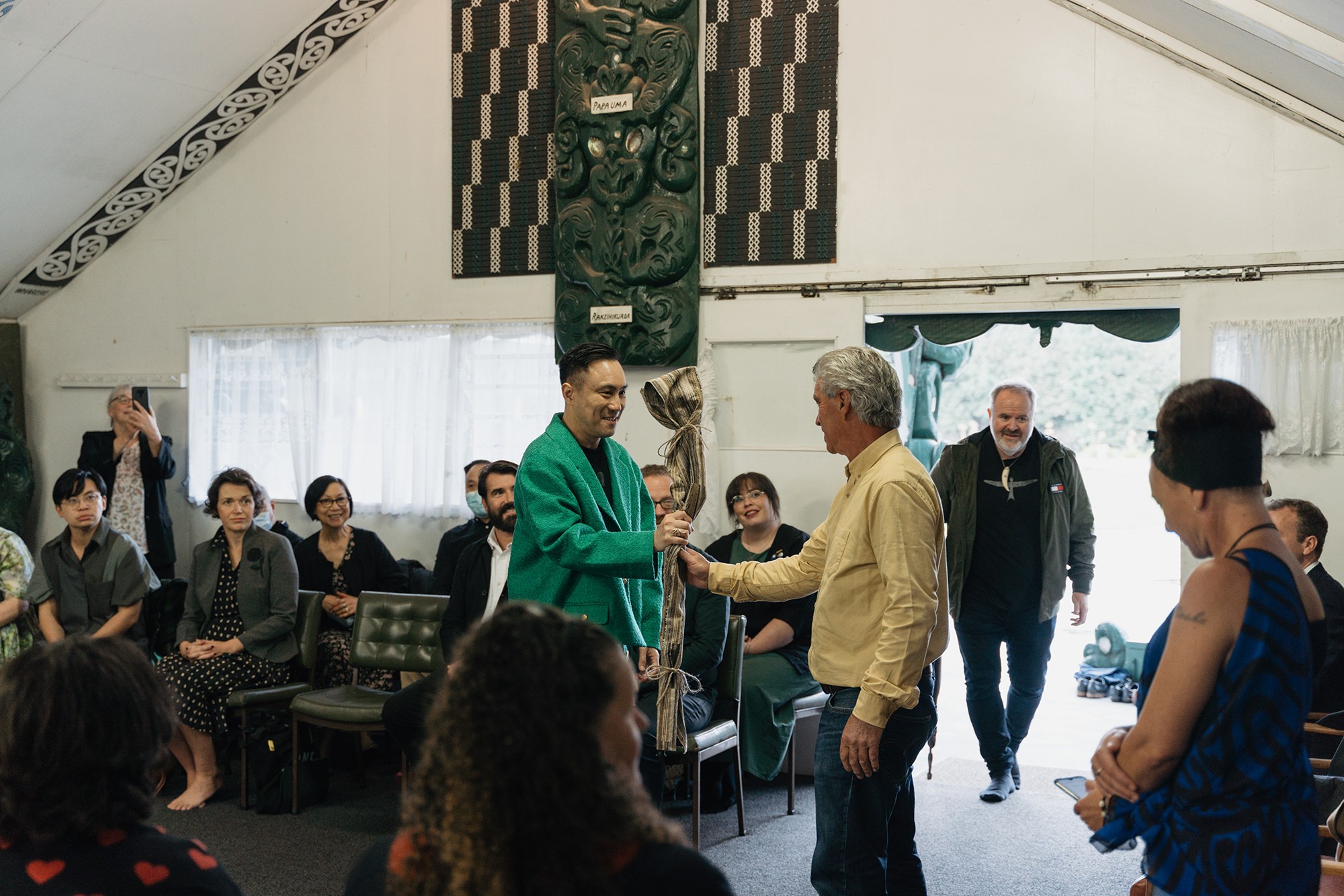 Inside a marae, carver Jacob Scott hands Chris Tse a tokotoko.