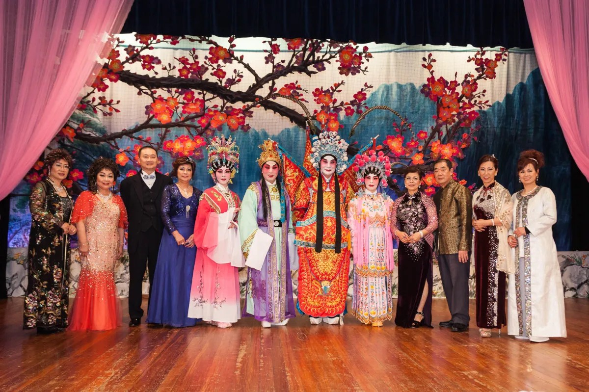 A group of performers in Cantonese opera costume stand on a stage in front of a blossom backdrop