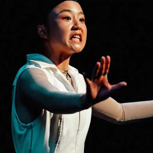 A woman dressed in a plain white puffer jacket and a black bald cap gestures in the air