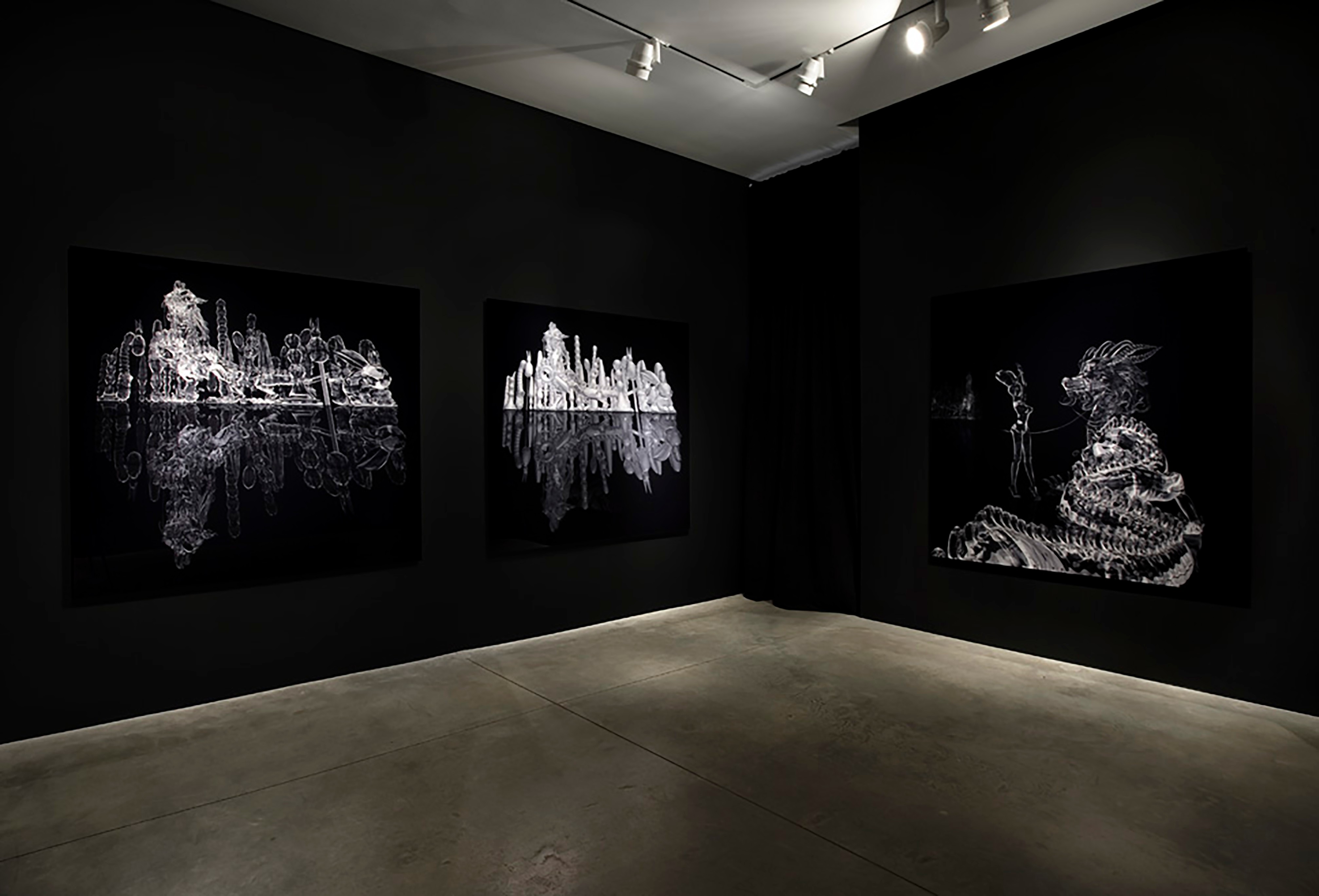 Three large black works with shiny white and silver figures in the corner of an art gallery with black walls.