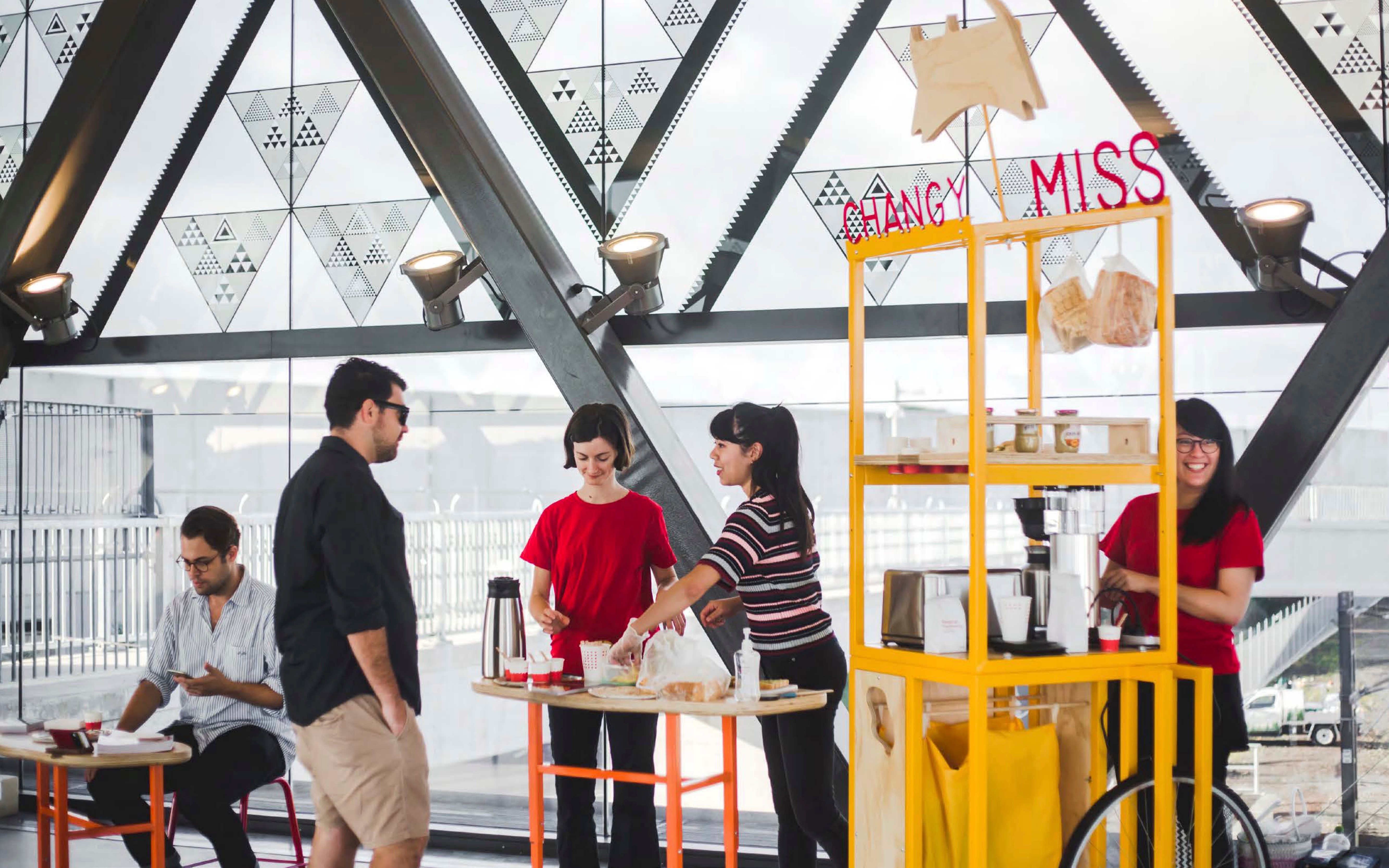 A yellow metal cart serving coffee next to orange and wooden tables where pastries are being served.