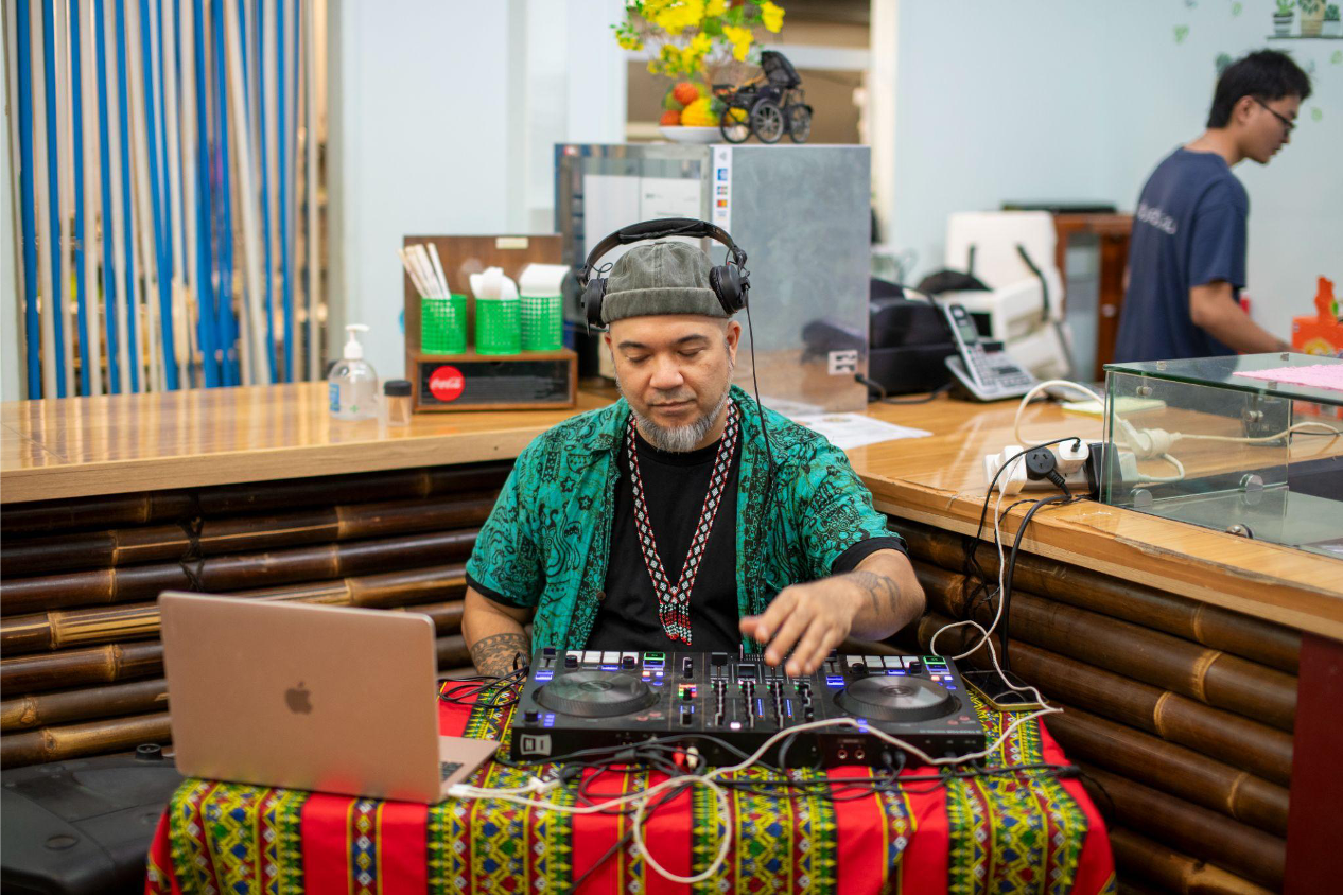 A guy in a colourful shirt sits at a small table DJing.