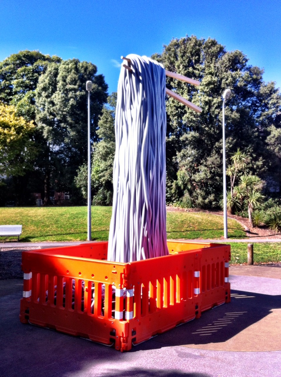 Orange roadwork barricades surrounding a tower of noodle-like objects being lifted with a giant pair of chopsticks by an invisible hand. In the background is a pathway with a bench seat, lampposts, and a grassy area with trees behind it. 