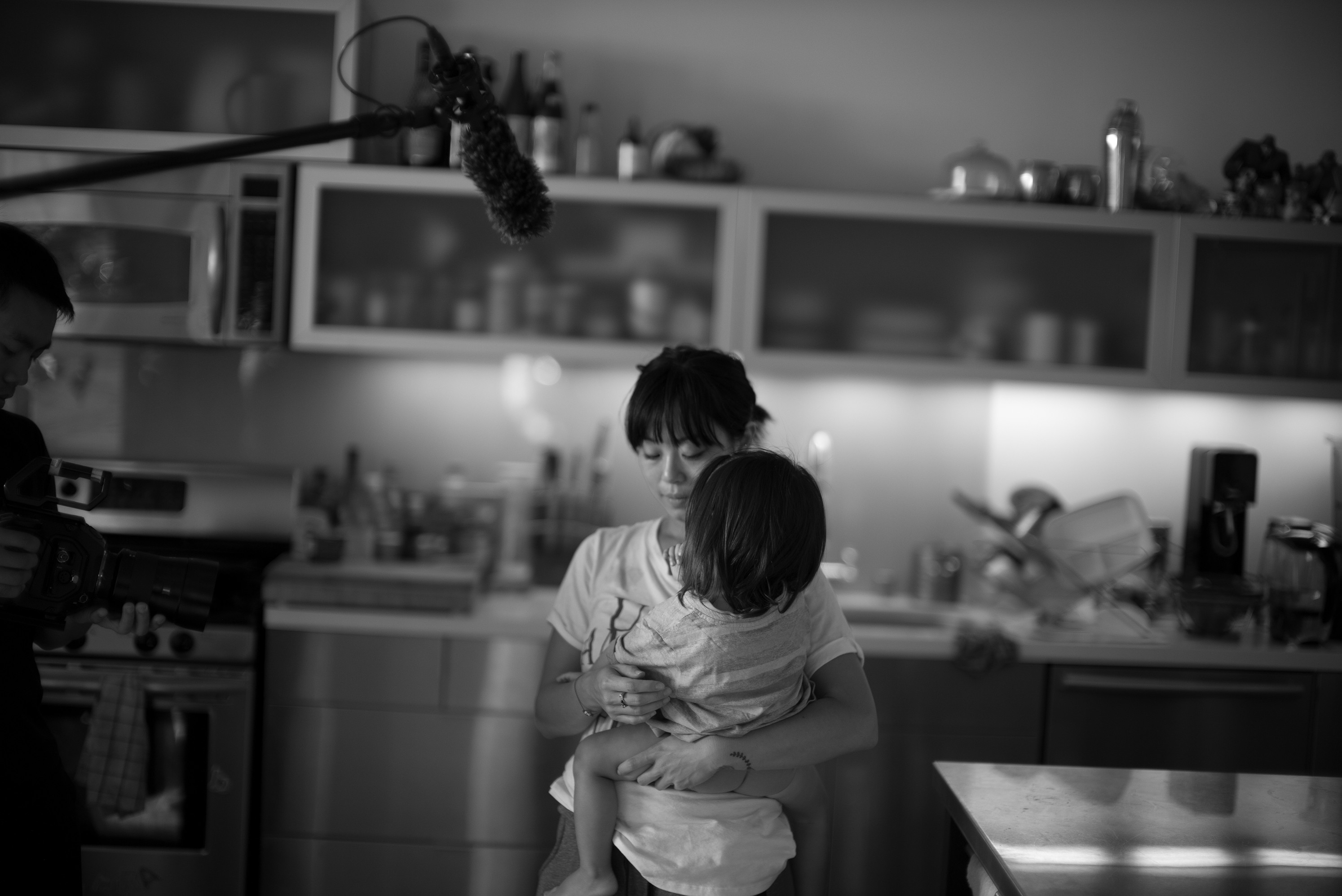 Black and white behind-the-scenes image of a film shoot in a kitchen, showing a woman holding a young child while a crew member films with a camera and a boom microphone hovers above.