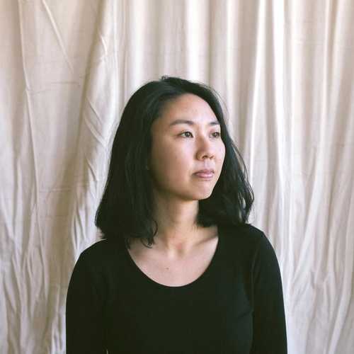 Photographic portrait of a woman with short black hair in front of a draped beige background. She is wearing black clothing and is looking to the side away from the camera.