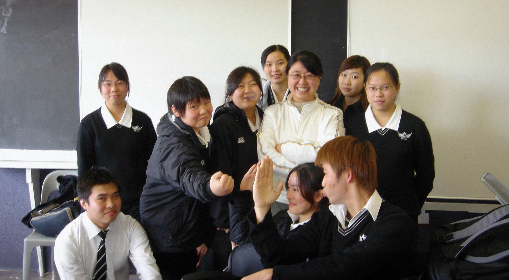 In a classroom, nine students in uniforms pose in a group with their teacher, standing in the centre.