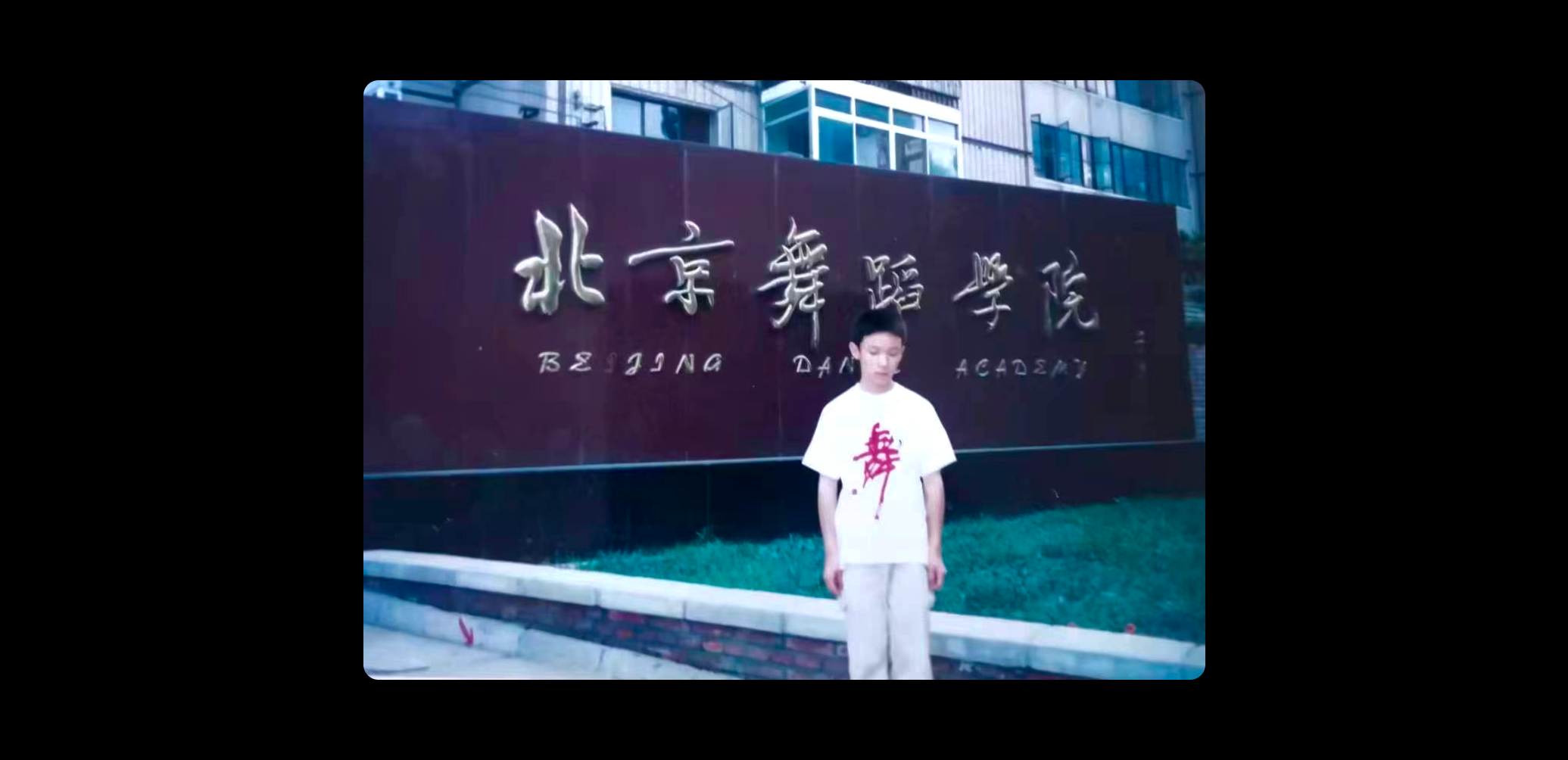 A young boy in a white t-shirt looks at the ground and stands in front of a Beijing Dance Academy sign.