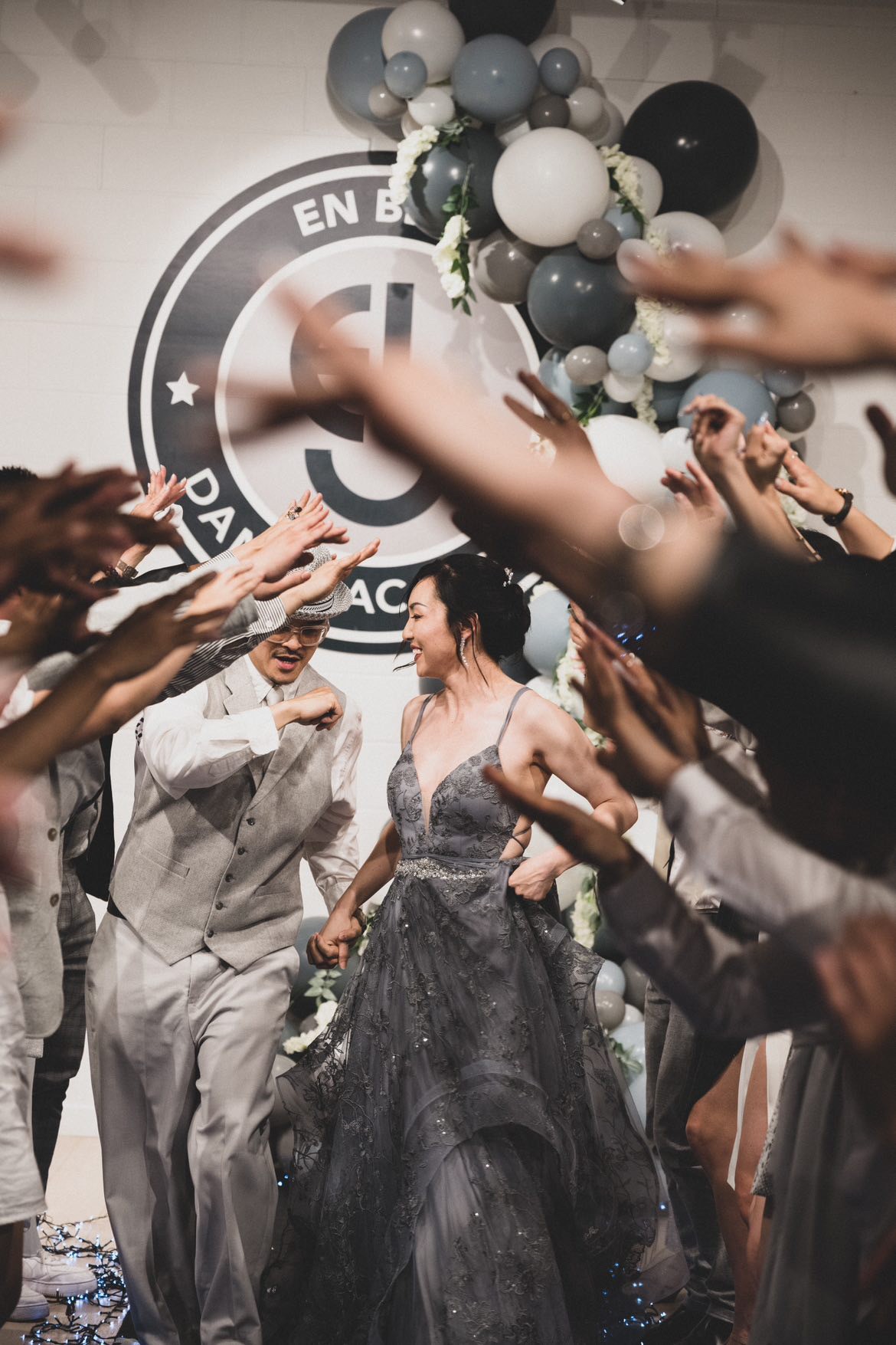 A man in a grey suit holding hands with a woman in a grey gown as they walk back down the aisle with everybody creating an archway with their arms