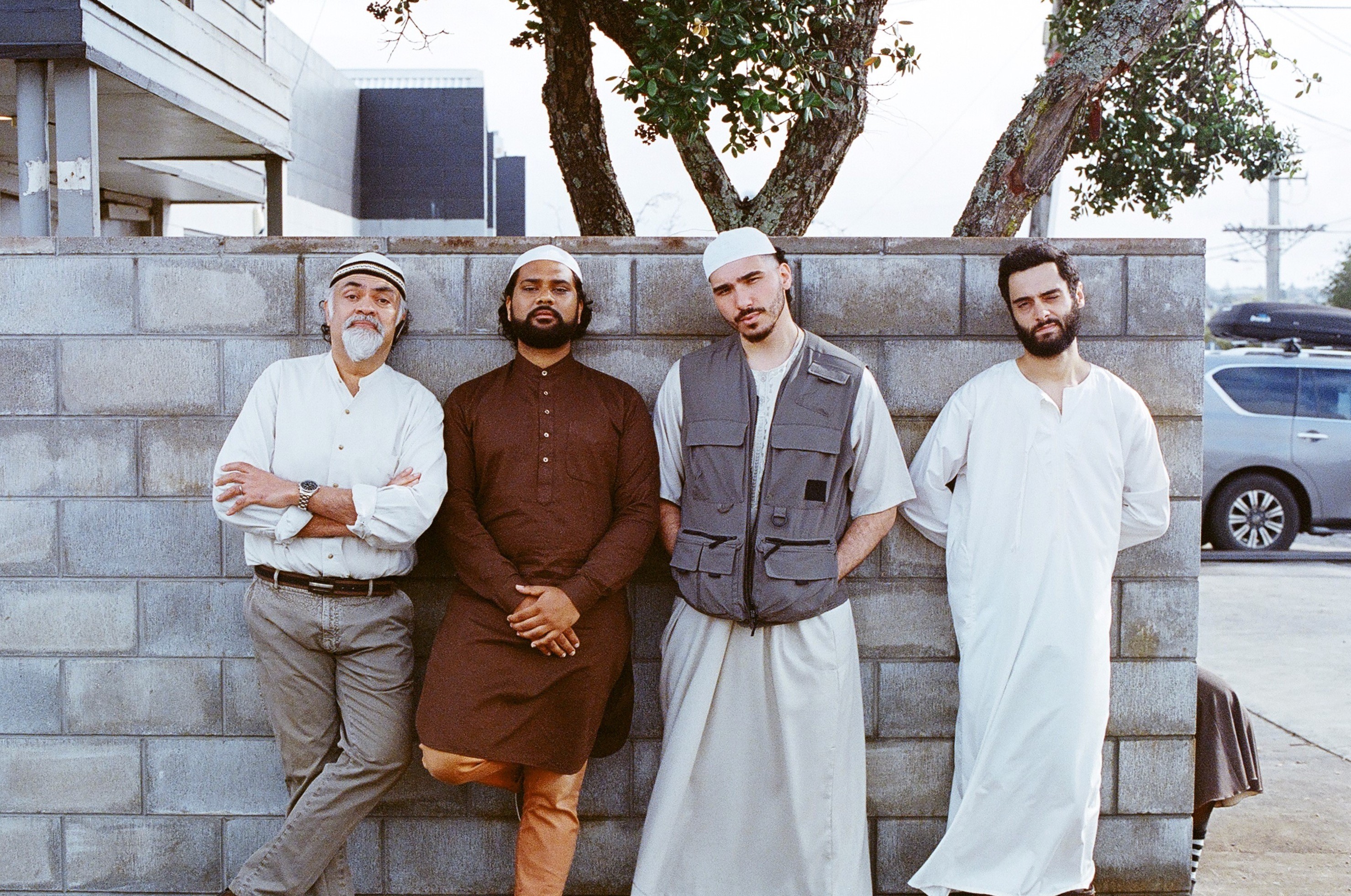 Four men in traditional and contemporary clothing stand against a concrete wall, posing casually on set outdoors.