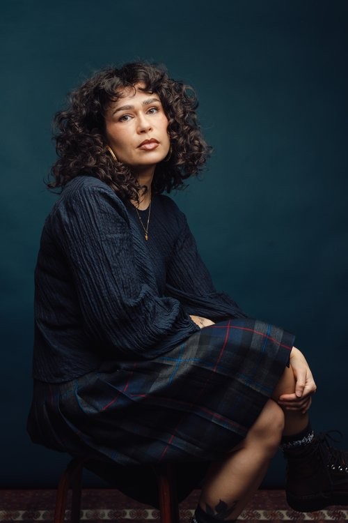 a woman with curly brown hair and a navy dress looks at the camera