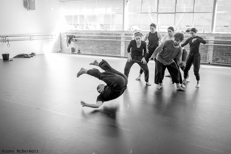 A group of dancers stand behind Xin performing a backwards leap in a rehearsal.