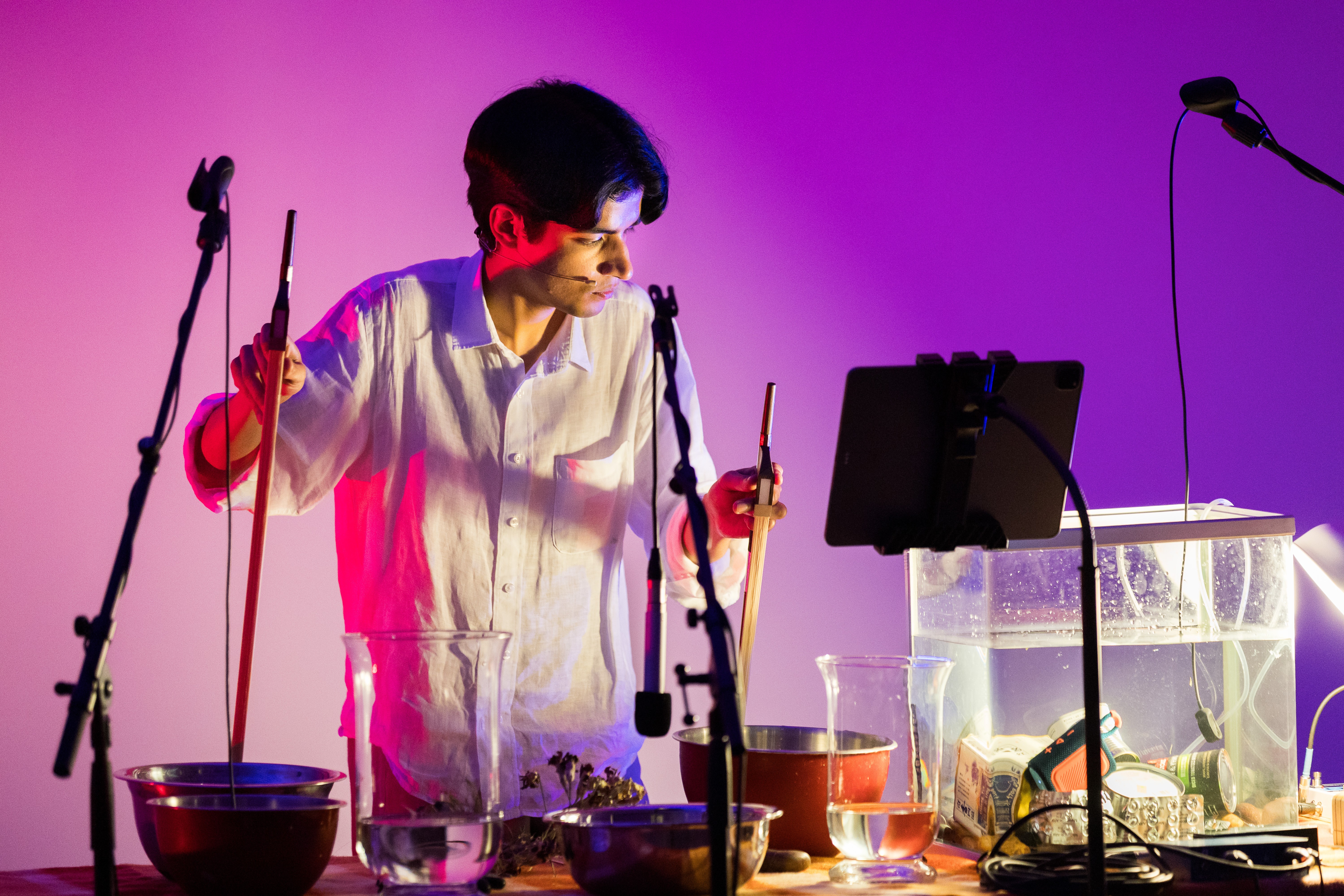 A South Asian performer holds two bowstrings to mixing bowls. He is standing at a table full of objects and concentrates on a musical score.