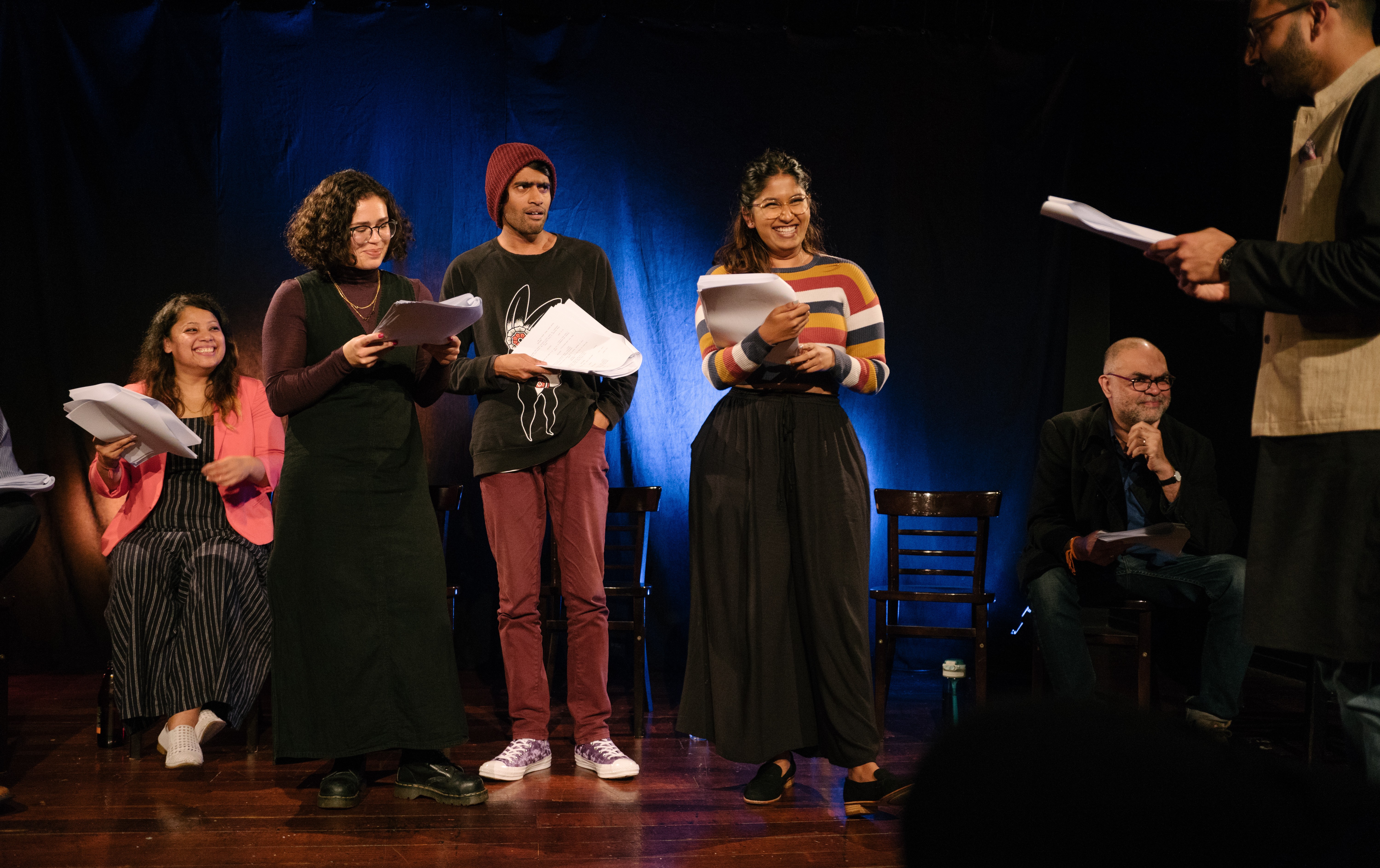 Three South Asian actors holding scripts stand in a group looking at an actor in the foreground. One of the actors smiles at him while another looks shocked. Two people sit behind the action on chairs waiting for their scene.