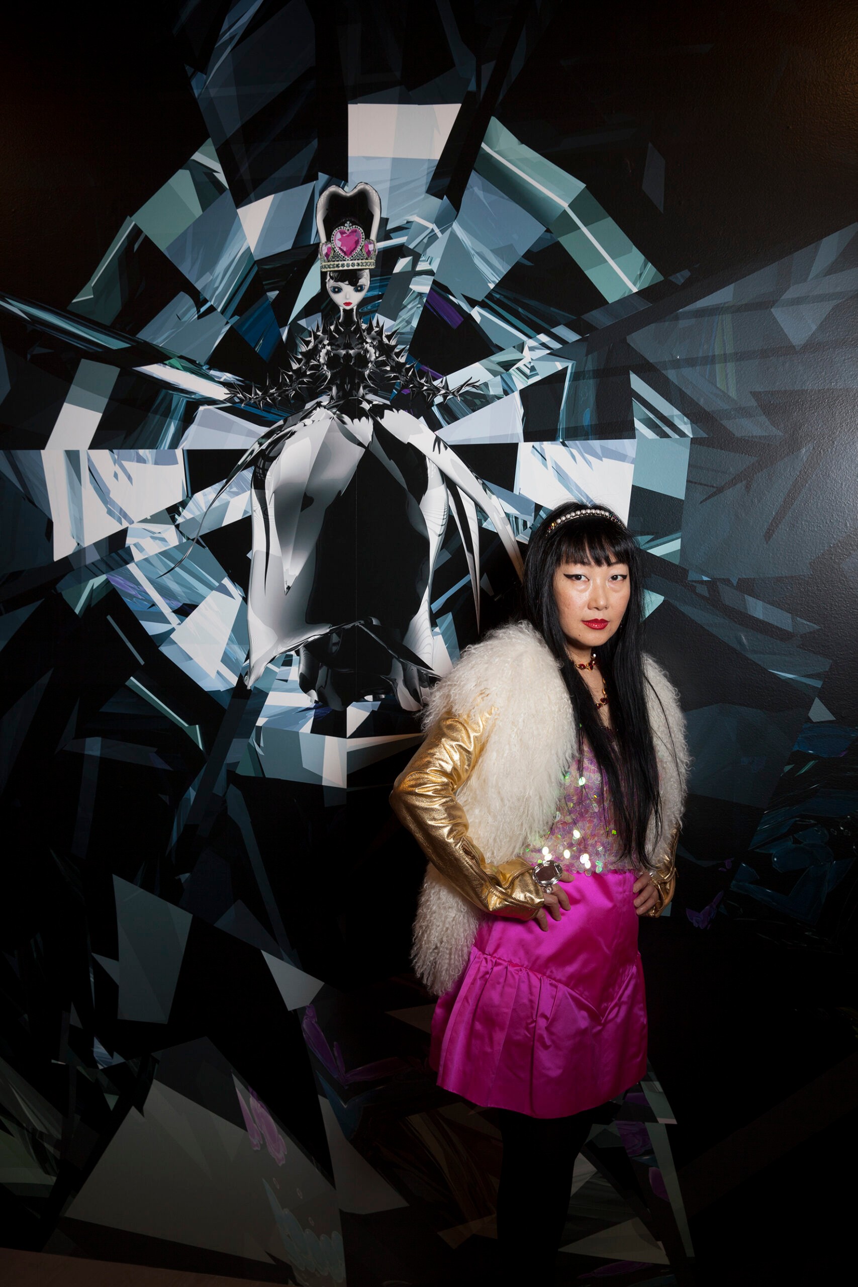 A woman with long dark hair poses in front of an artwork with a crystal design. She is wearing a hot pink dress, gold gloves, fur and a sparkly headband.