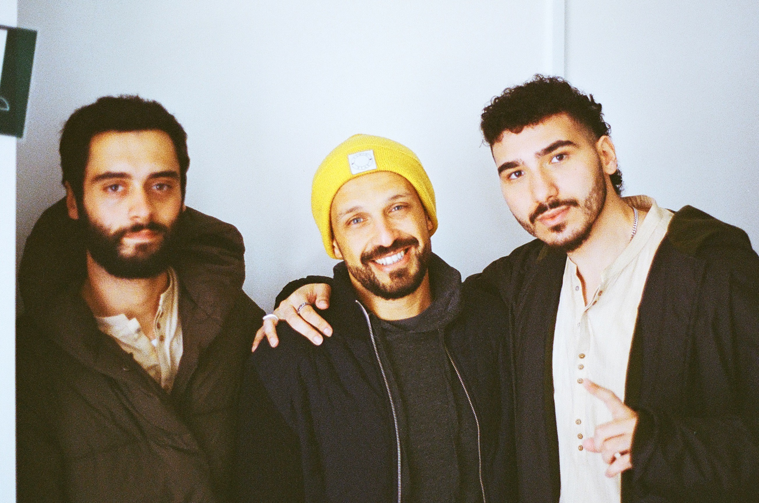 Three men stand close together, smiling at the camera; the man in the middle wears a yellow beanie.