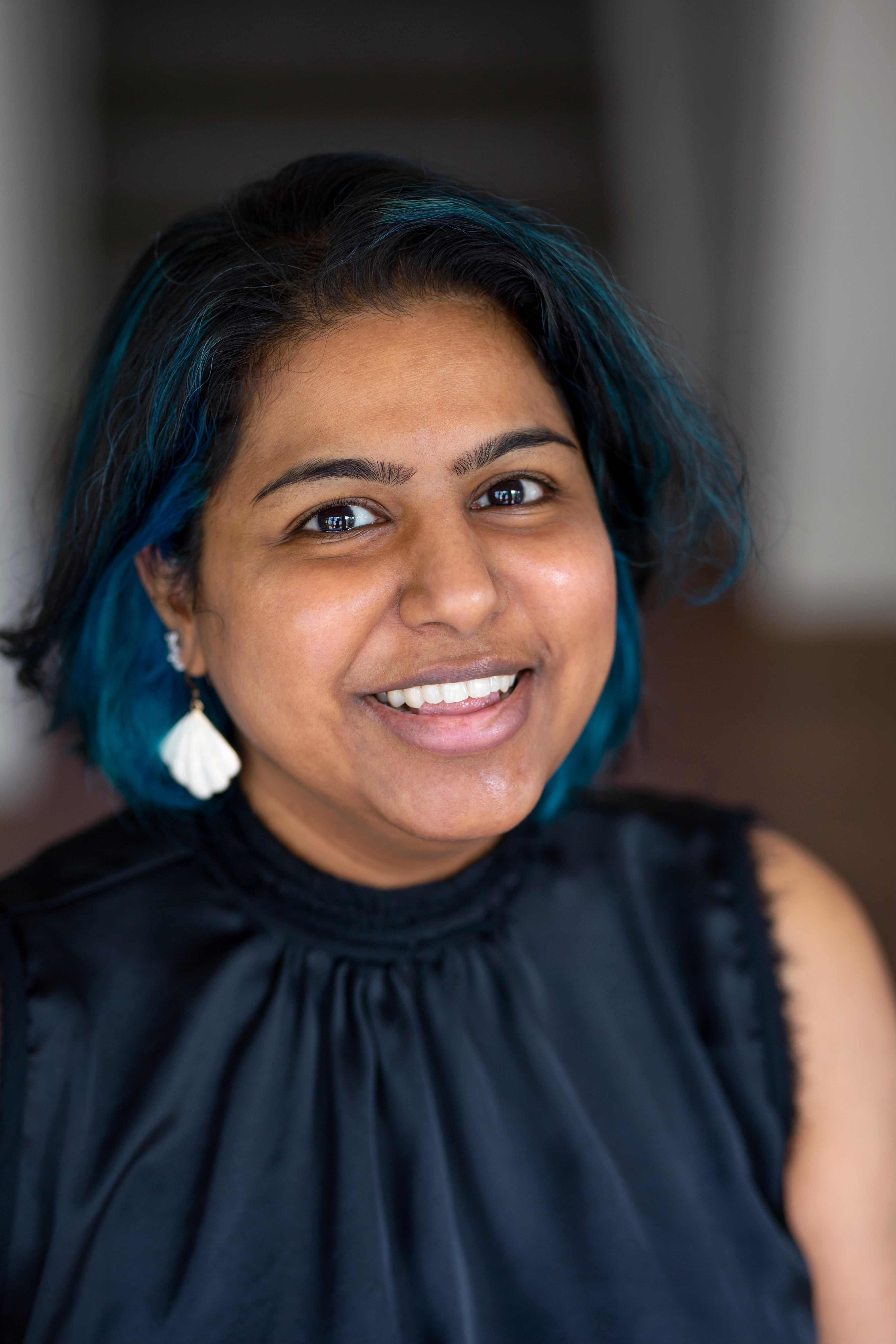 Close-up of a woman with a blue-black bob and white shell earrings smiling at the camera