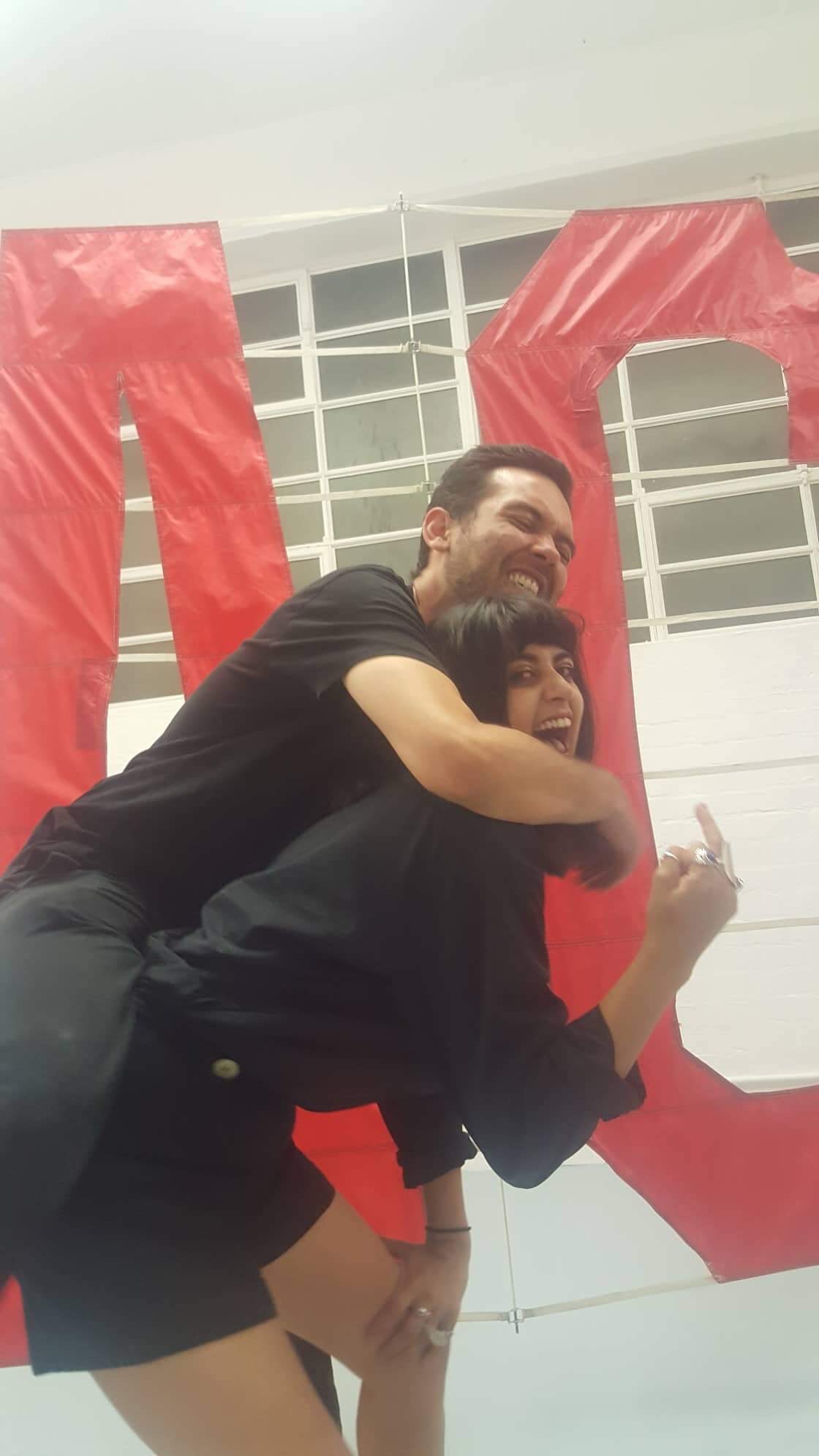 Photograph of two smiling people in black clothing. One person has climbed on the other person's back and is being carried by them. 