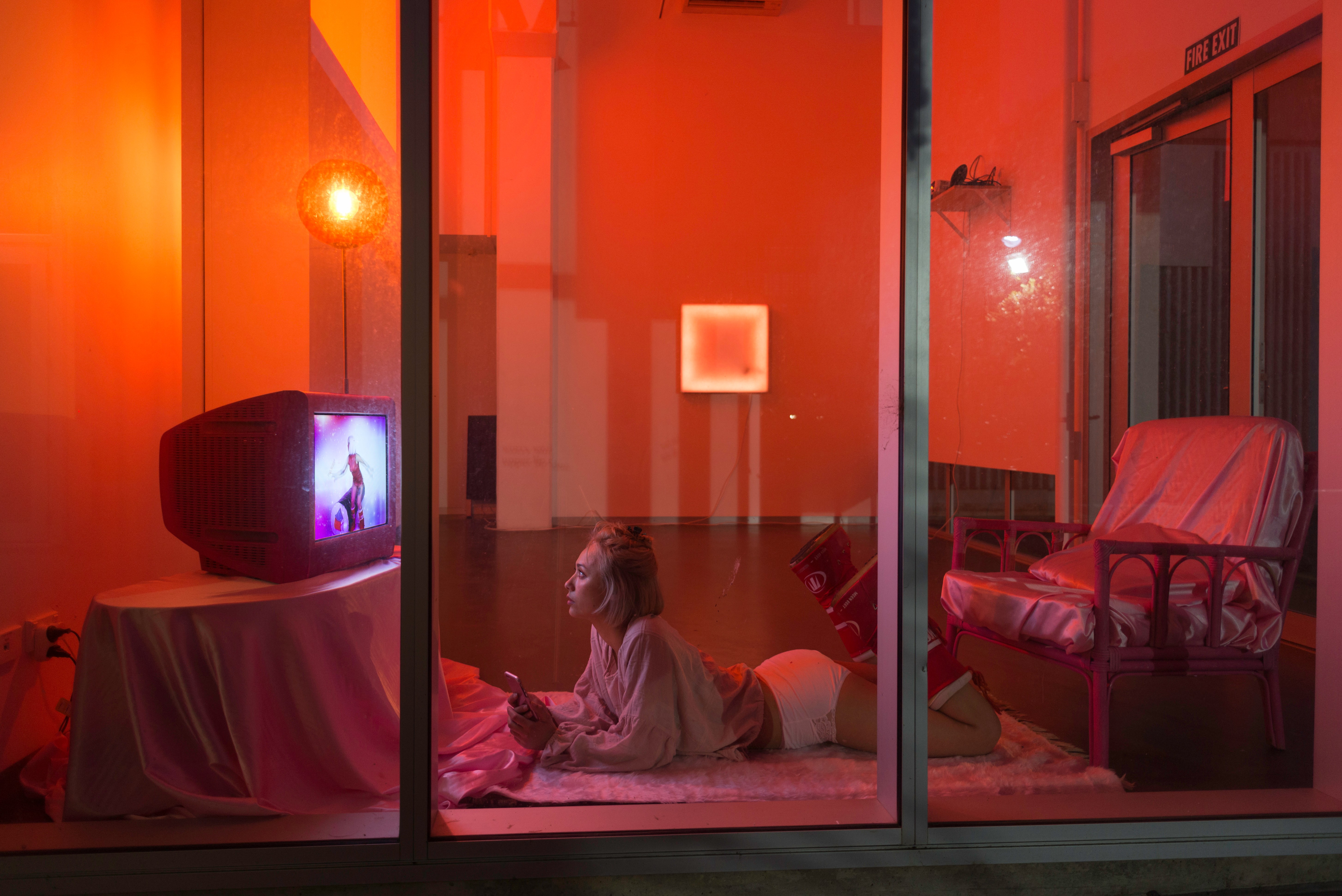 Bathed in orange light, Emiko Sheehan watches herself on a TV while lying on a rug.