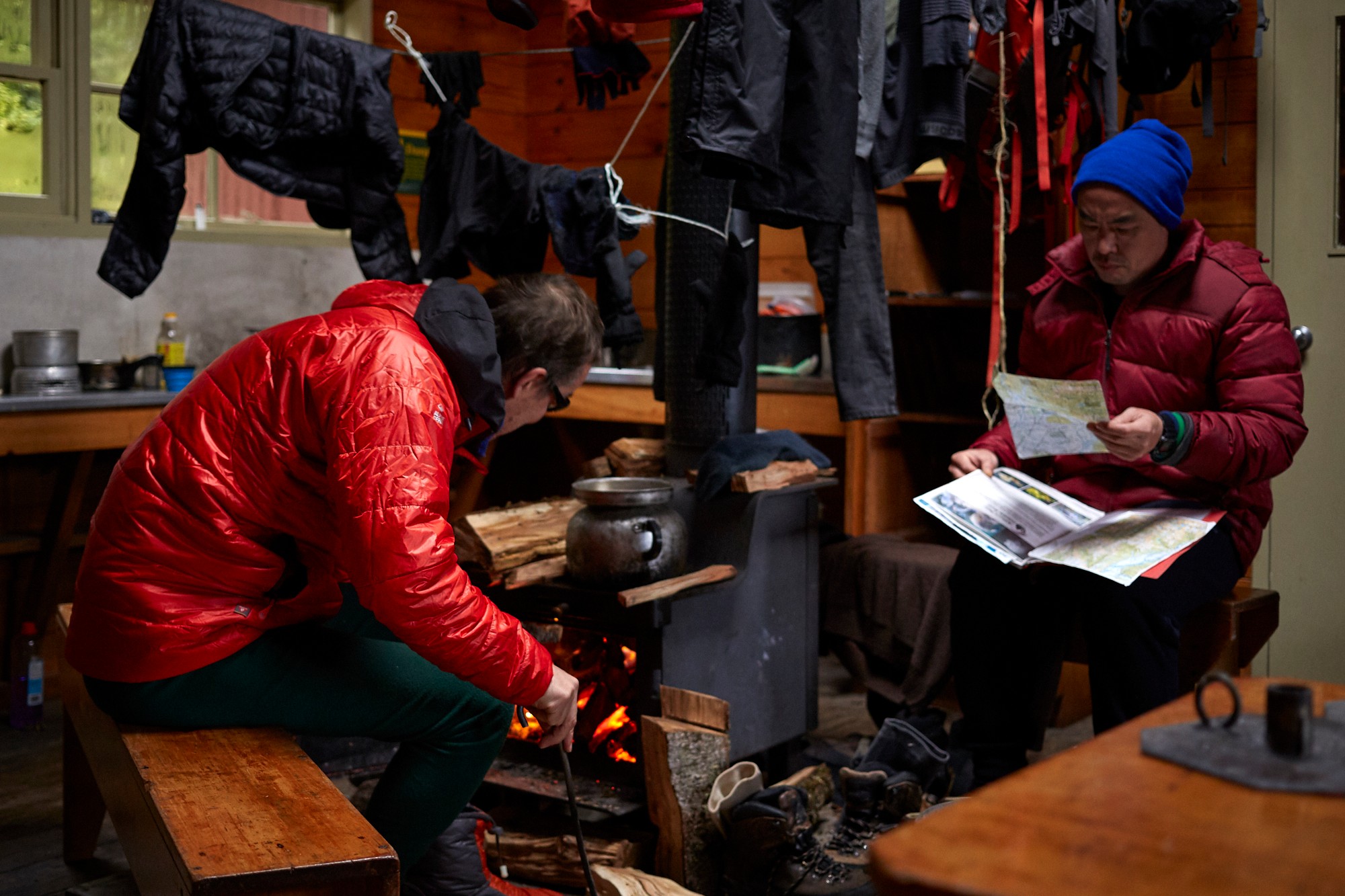 Two men in tramping gear sit in a hut while their things dry around them and they look at information.