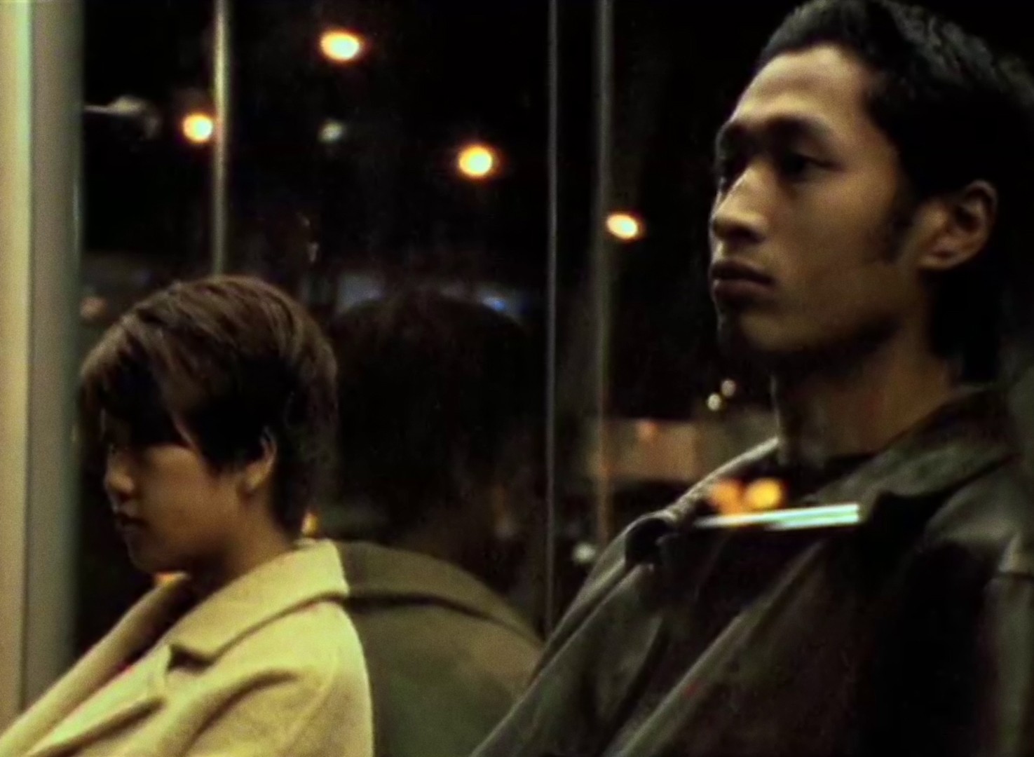 A woman wearing a beige overcoat and a man wearing a black leather jacket sit together at a bus stop.