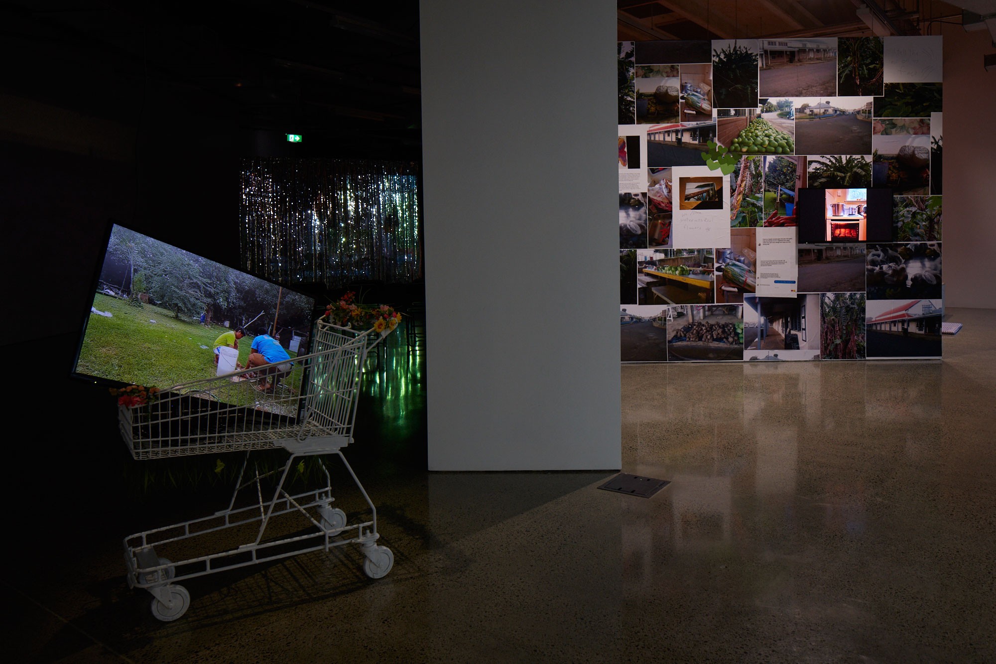 A TV screen with a video work is in a supermarket trolley in front of a shiny curtain and a wall covered in photos.