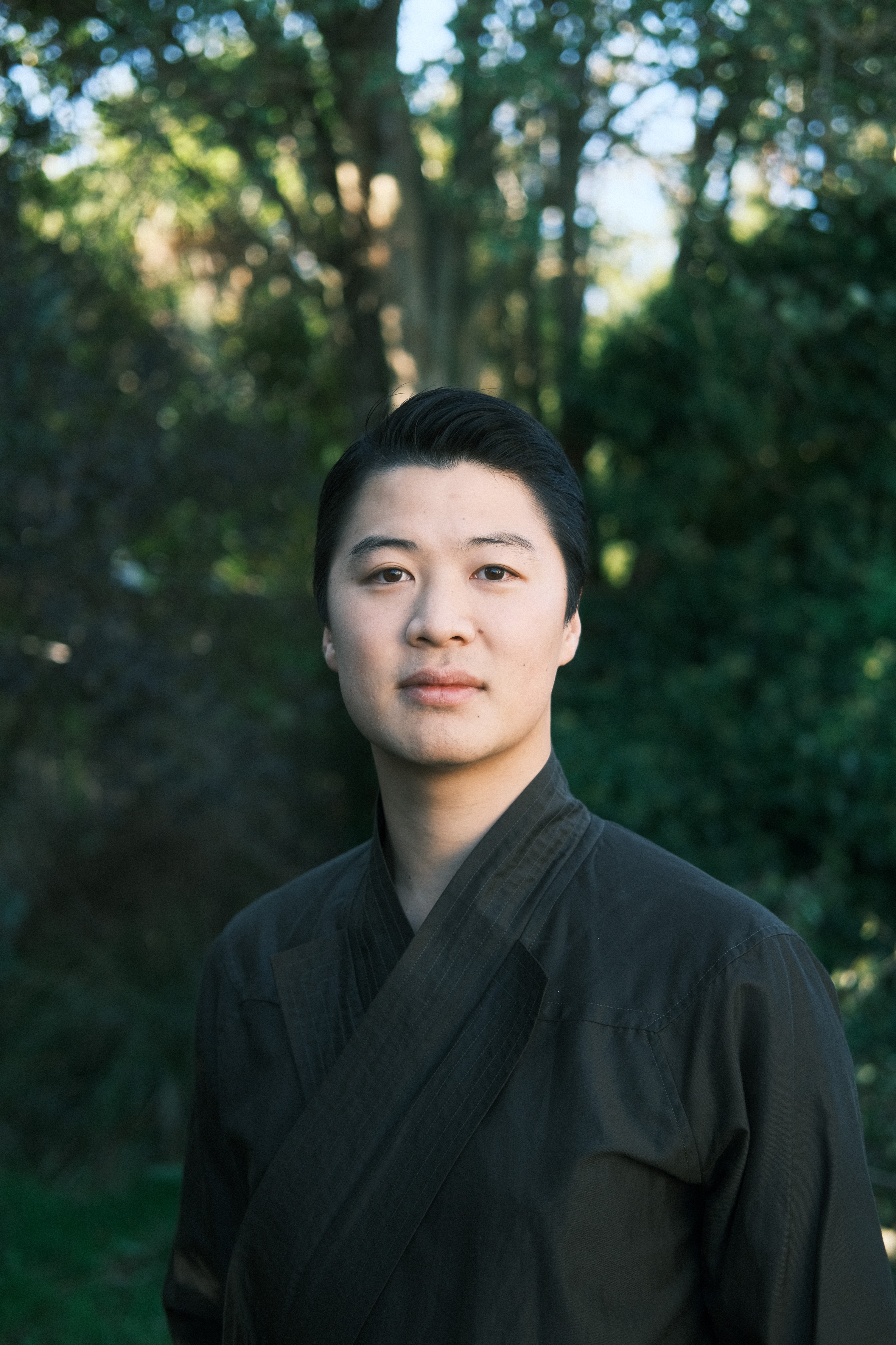 A man with short black hair wearing a dark wrap top, standing in front of trees