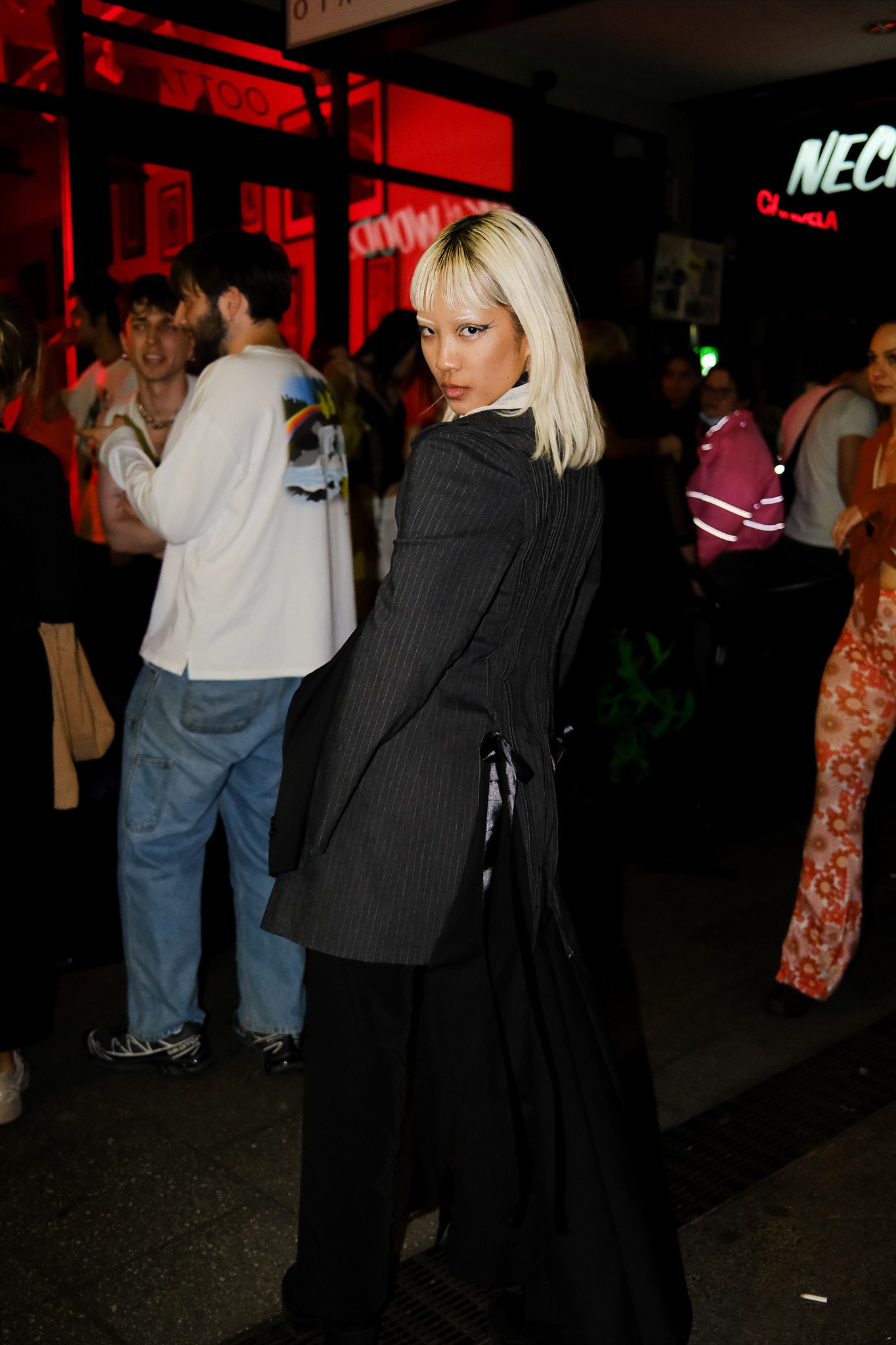 A person with bleached blonde hair and eyebrows in a stylish grey pinstripe jacket and black pants posing in front of a crowd of evening party goers