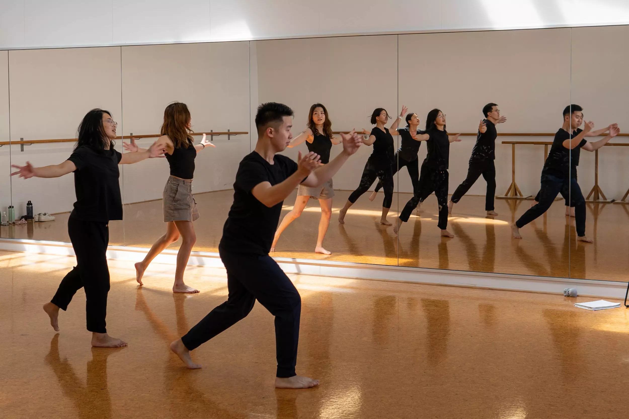 Three Asian dancers, dressed mostly in black rehearsal clothes, practising in a mirrored dance studio.