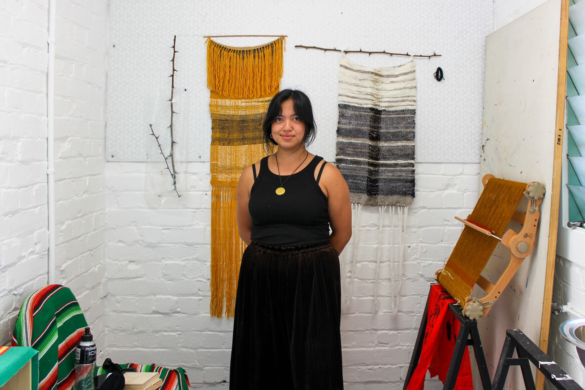 Person of Filipino descent in a black dress standing in front of two woven art pieces and a loom