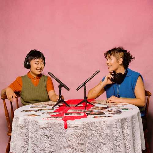 Two women sit at a lace-covered table in front of a pink wall. Microphones sit on the table along with photos spread across it. They each wear headphones.