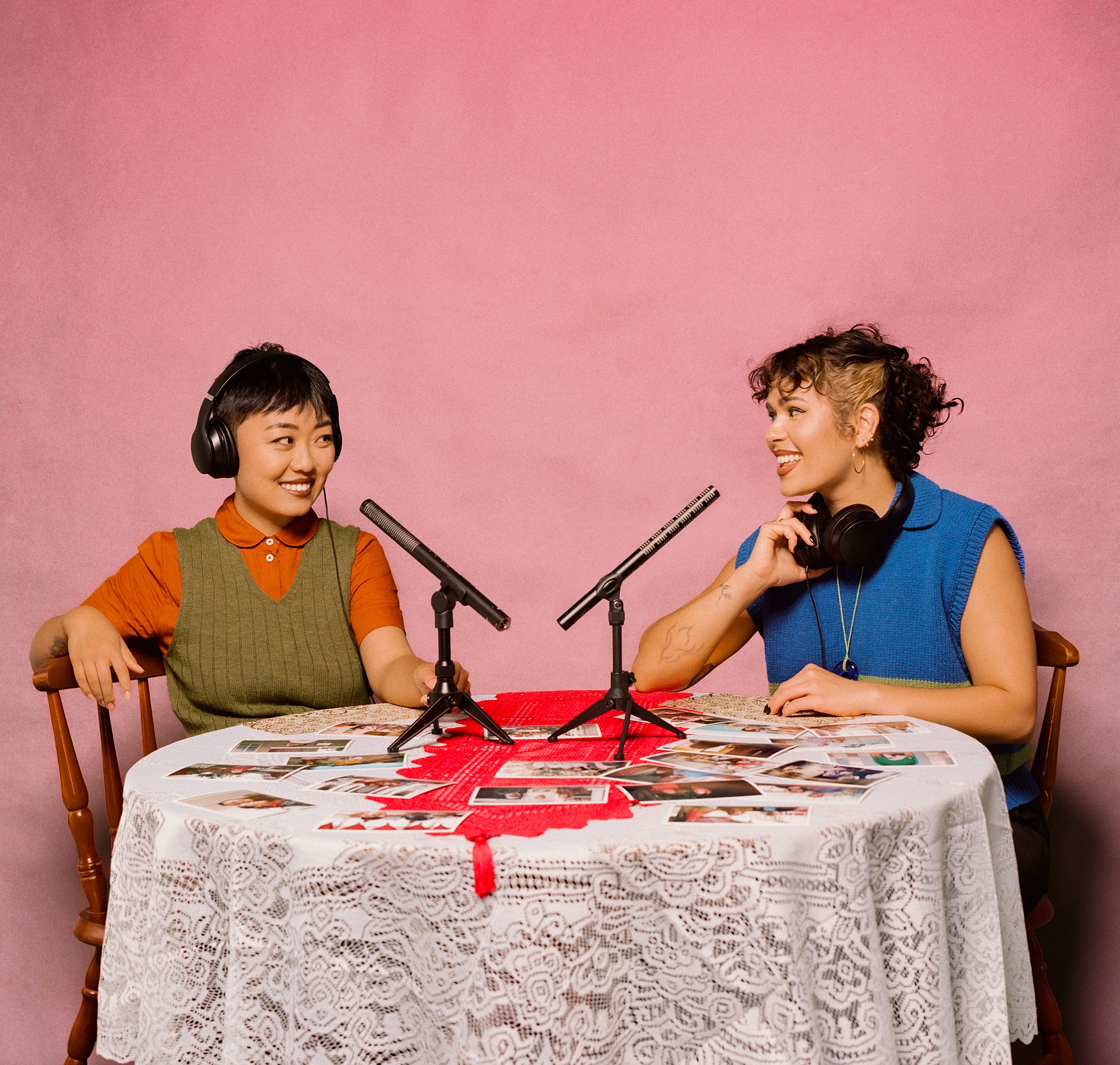 Two women sit at a lace-covered table in front of a pink wall. Microphones sit on the table along with photos spread across it. They each wear headphones.