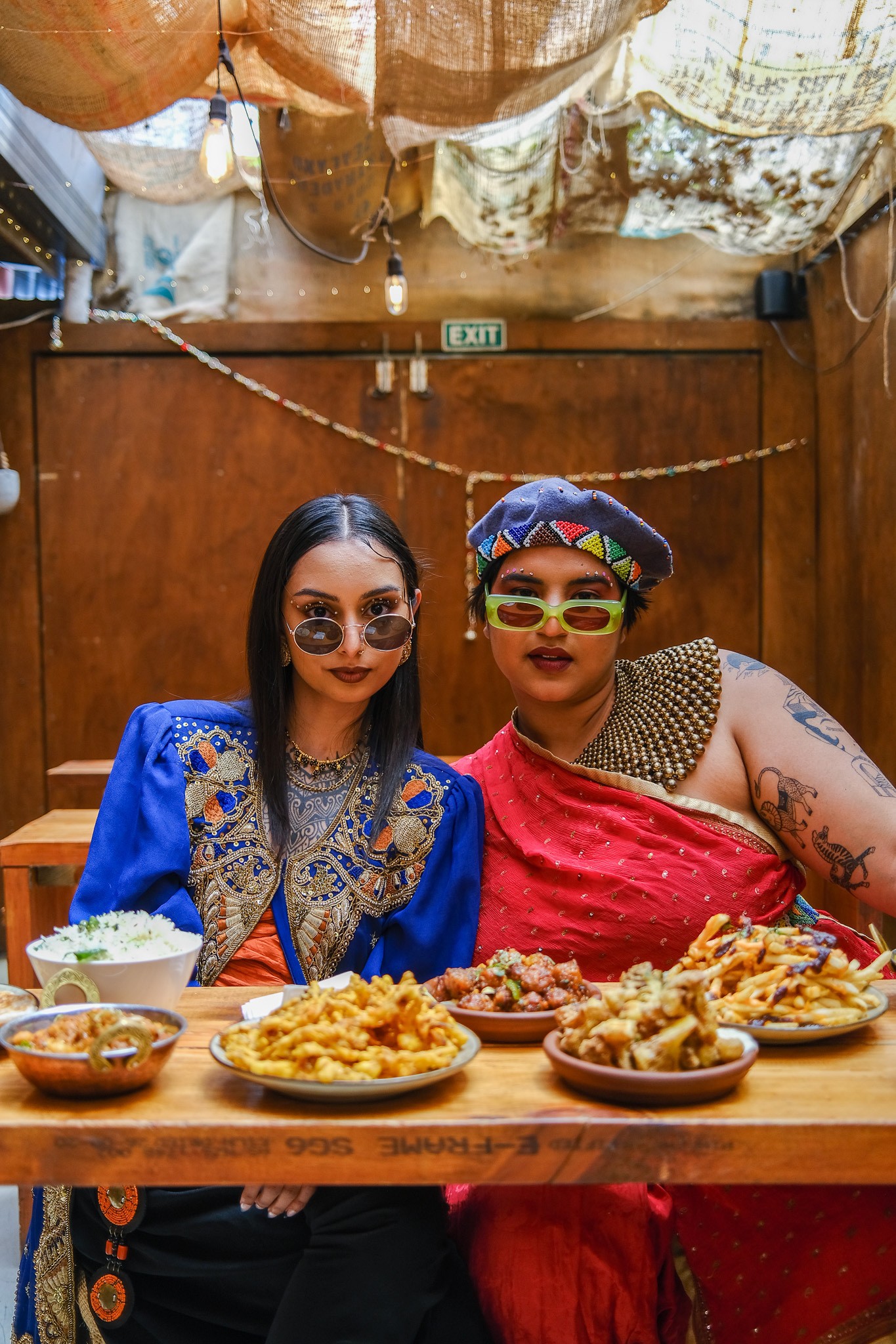 Two people of South Asian descent, wearing colourful South Asian-inspired outfits seated in front of a spread of South Asian food placed on a wooden table