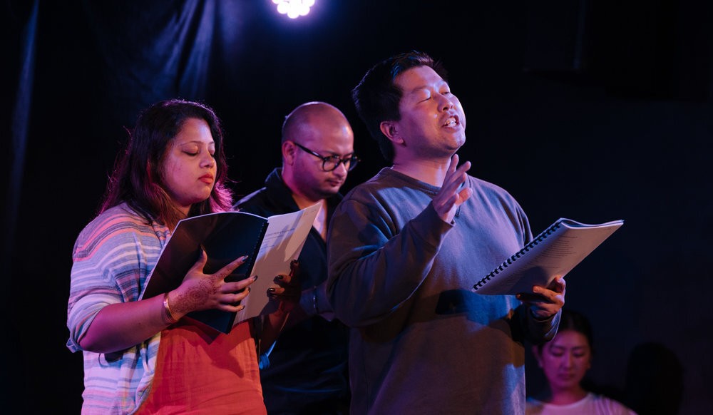 Three actors holding scripts read aloud.