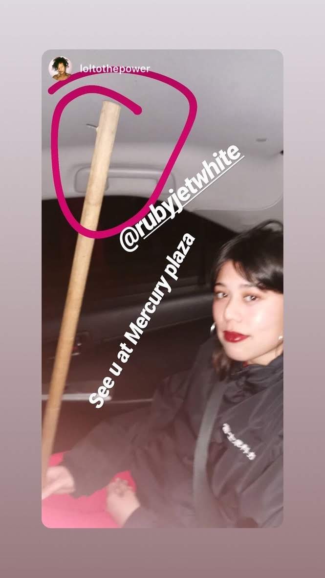 A woman with red lipstick in the back of a taxi, holding a wooden stick with a nail in it