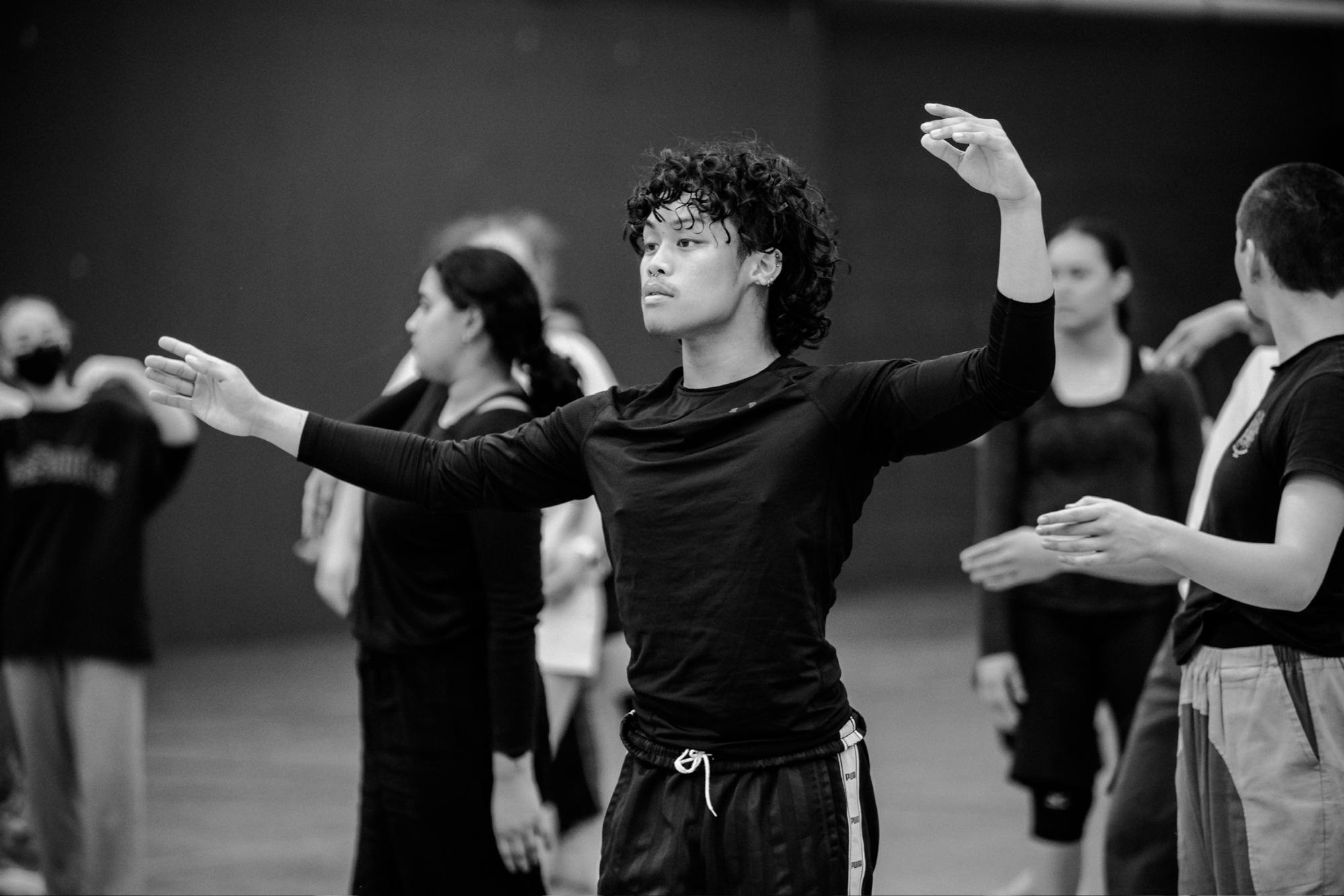 Black and white photo of Filipino-looking person with curly hair wearing black long sleeves doing dance rehearsals with other people in the background