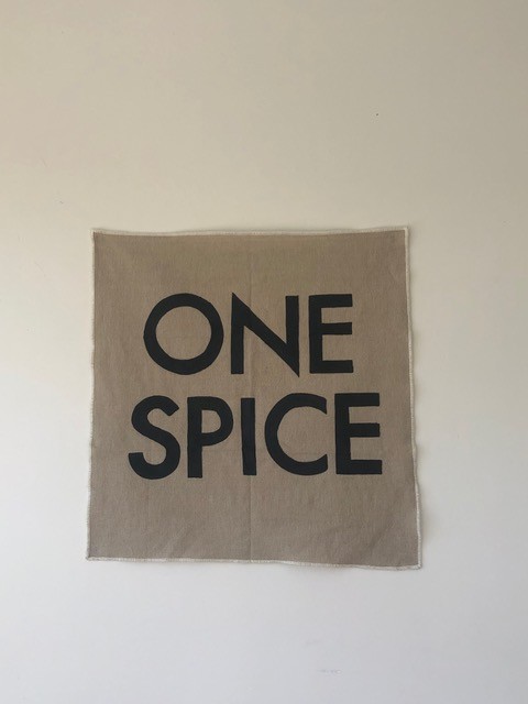 A piece of square textile fabric with the phrase 'ONE SPICE' printed in the centre