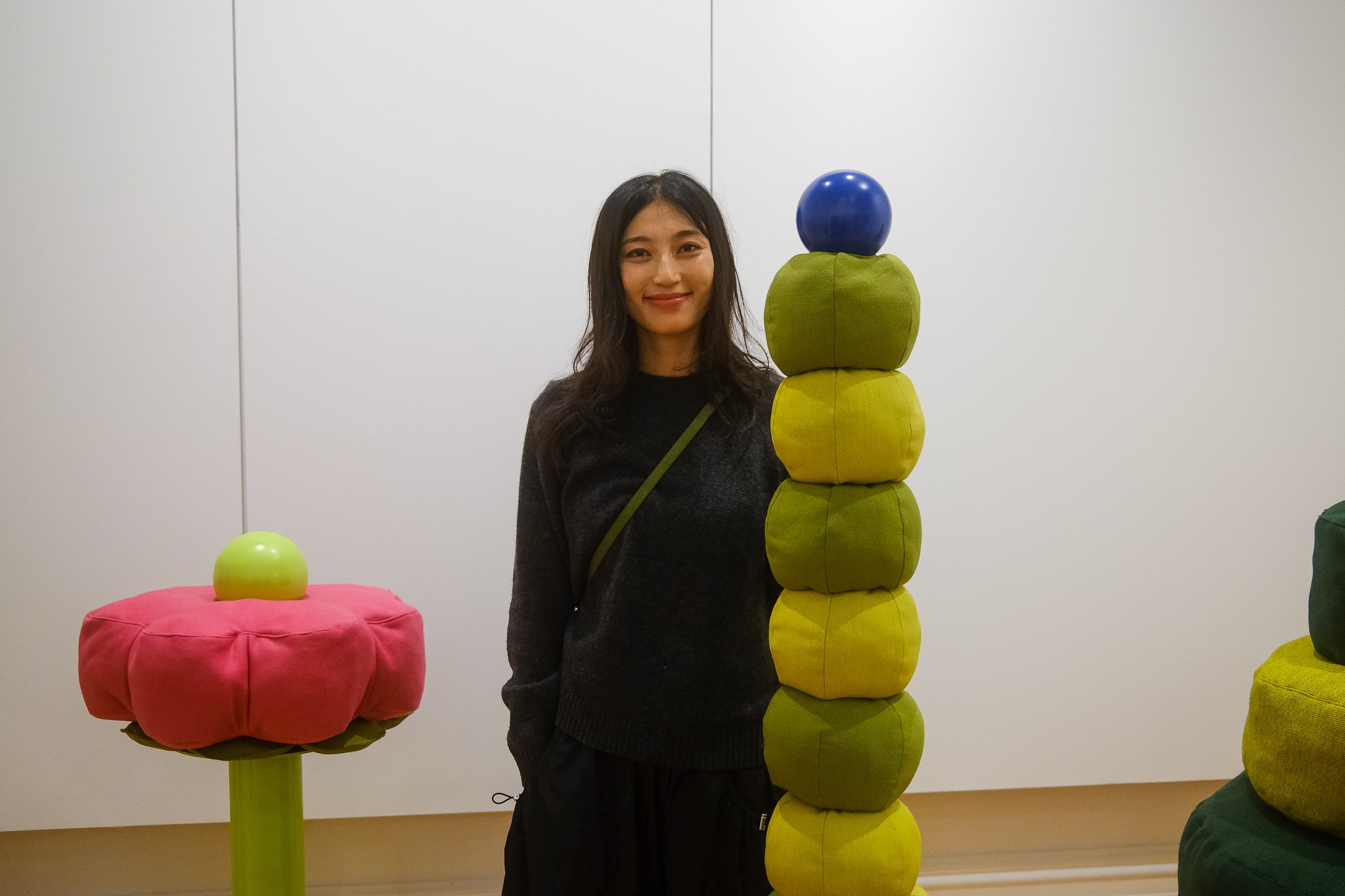 A woman in a black dress stands smiling behind a collection of brightly coloured soft sculptures evoking a garden