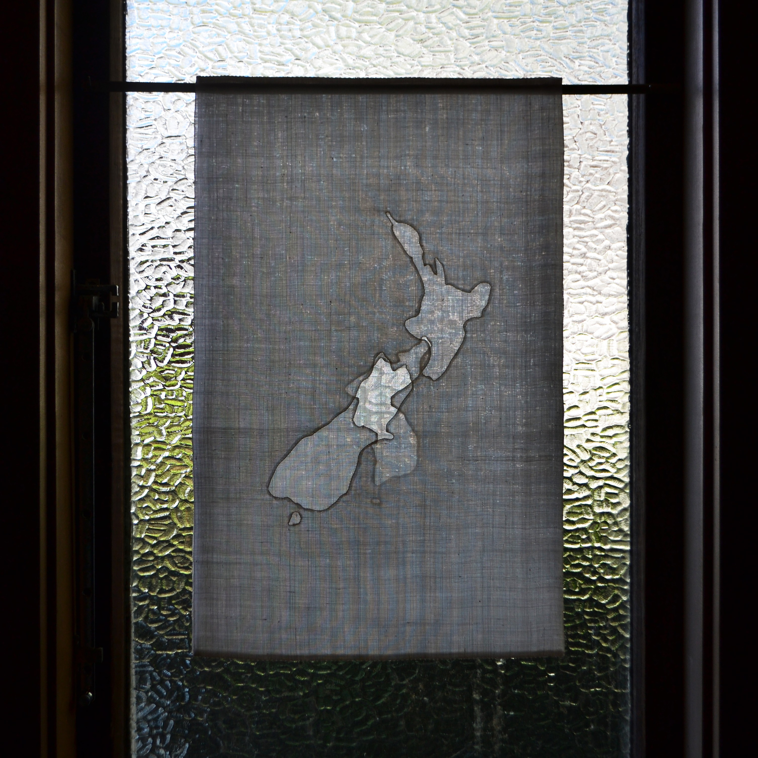 A fabric hanging with outlines of Korea and Aotearoa sewn into it, hanging in front of a window.