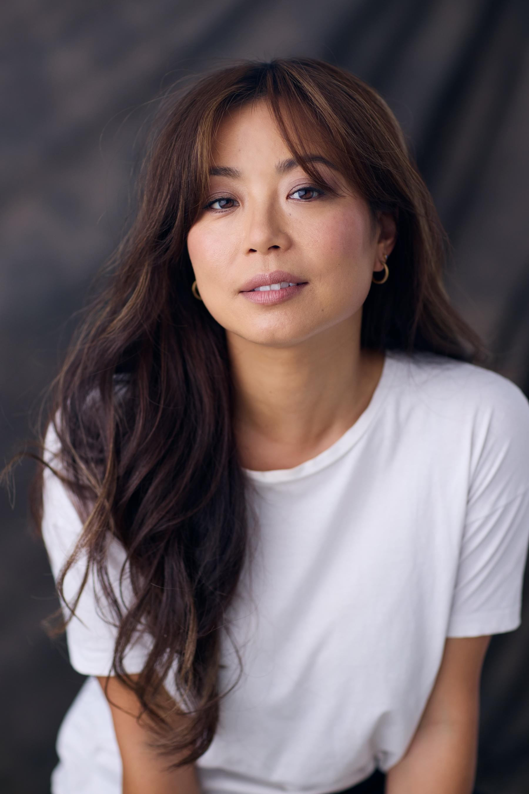 A woman with long brown hair and hoop earrings poses in a white t-shirt against a dark, softly blurred background.